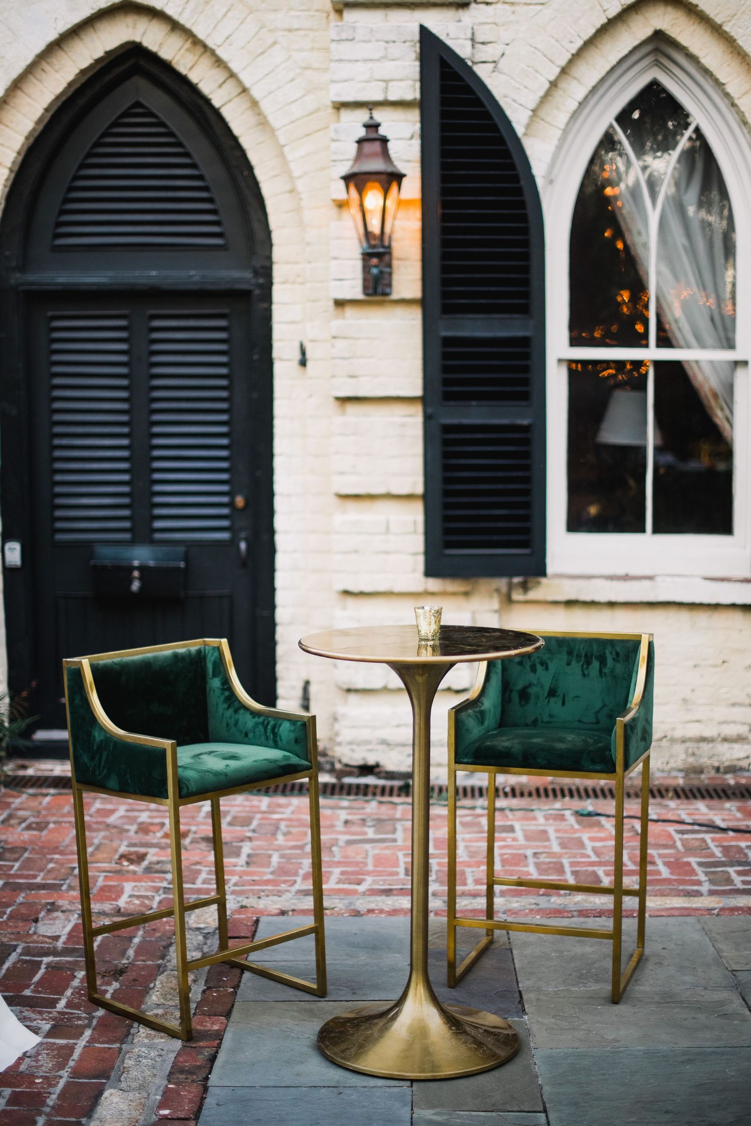 Elegant emerald green velvet chairs with gold frames arranged on a brick patio beneath Gothic arched windows.