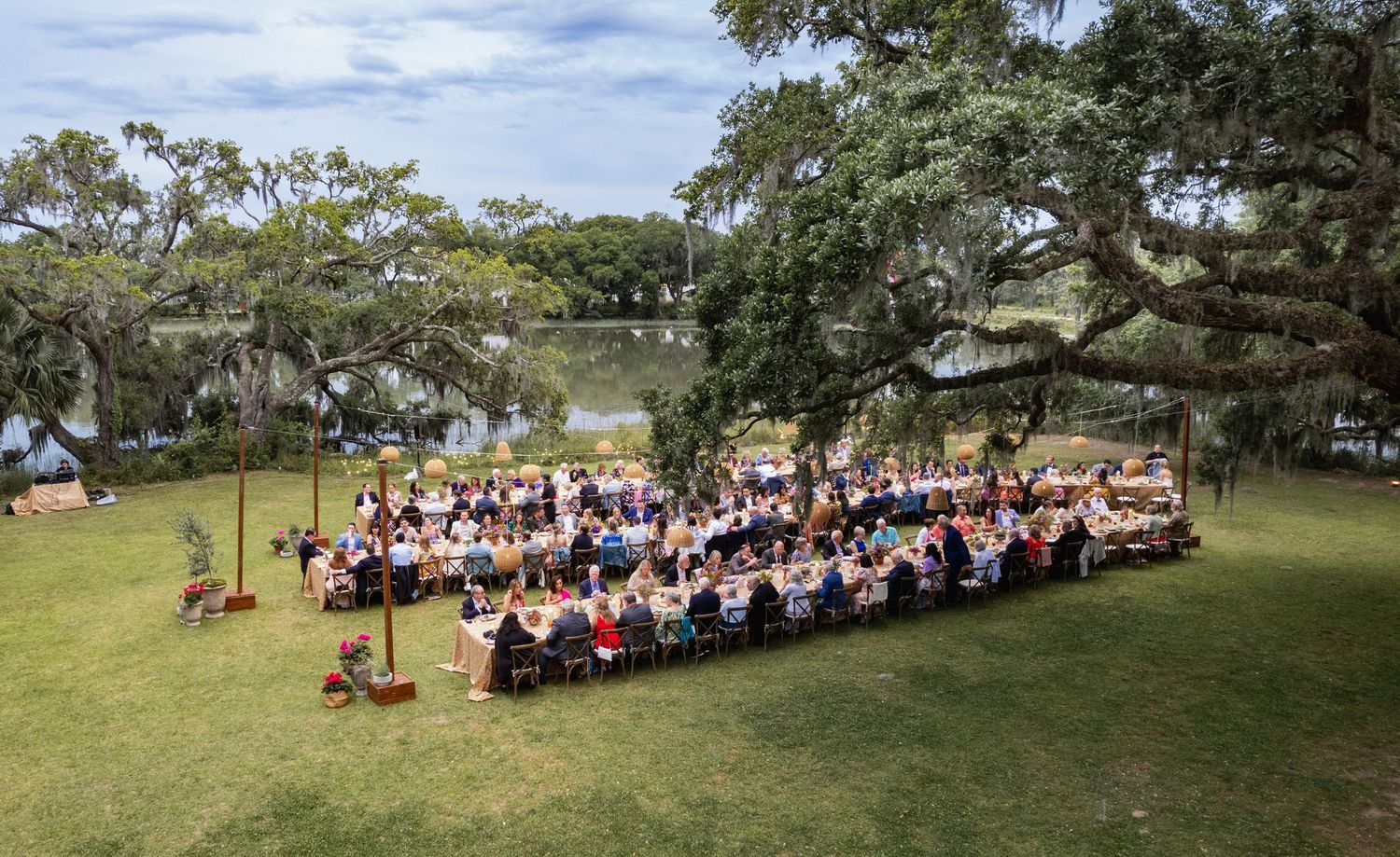 A panoramic view of a large outdoor gathering with people seated on chairs spread across a grassy lawn under trees.