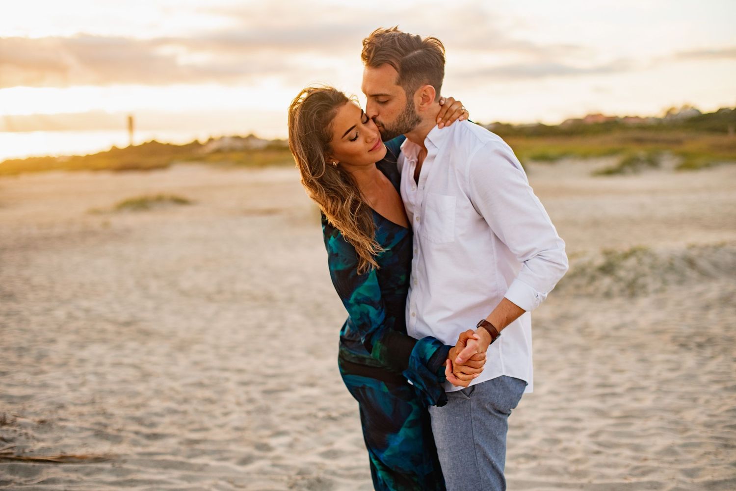 Romantic couple embraces on a sandy beach at sunset with soft golden light illuminating the scene.