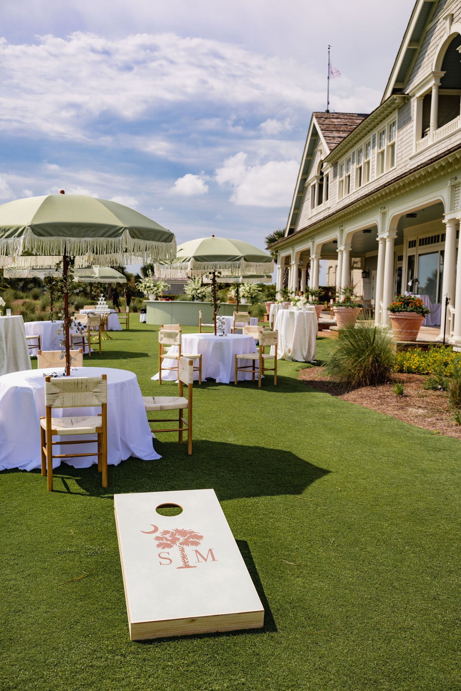 Outdoor wedding venue setup with cornhole games and white chairs on a manicured green lawn.