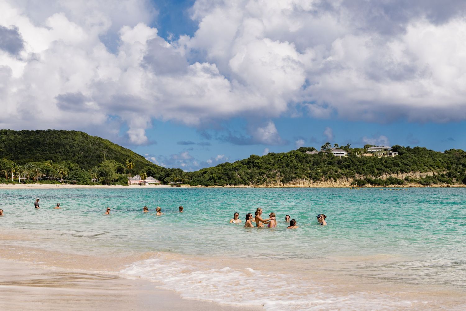 Beachgoers enjoy crystal clear turquoise waters at a tropical beach surrounded by lush green hills and white sand.