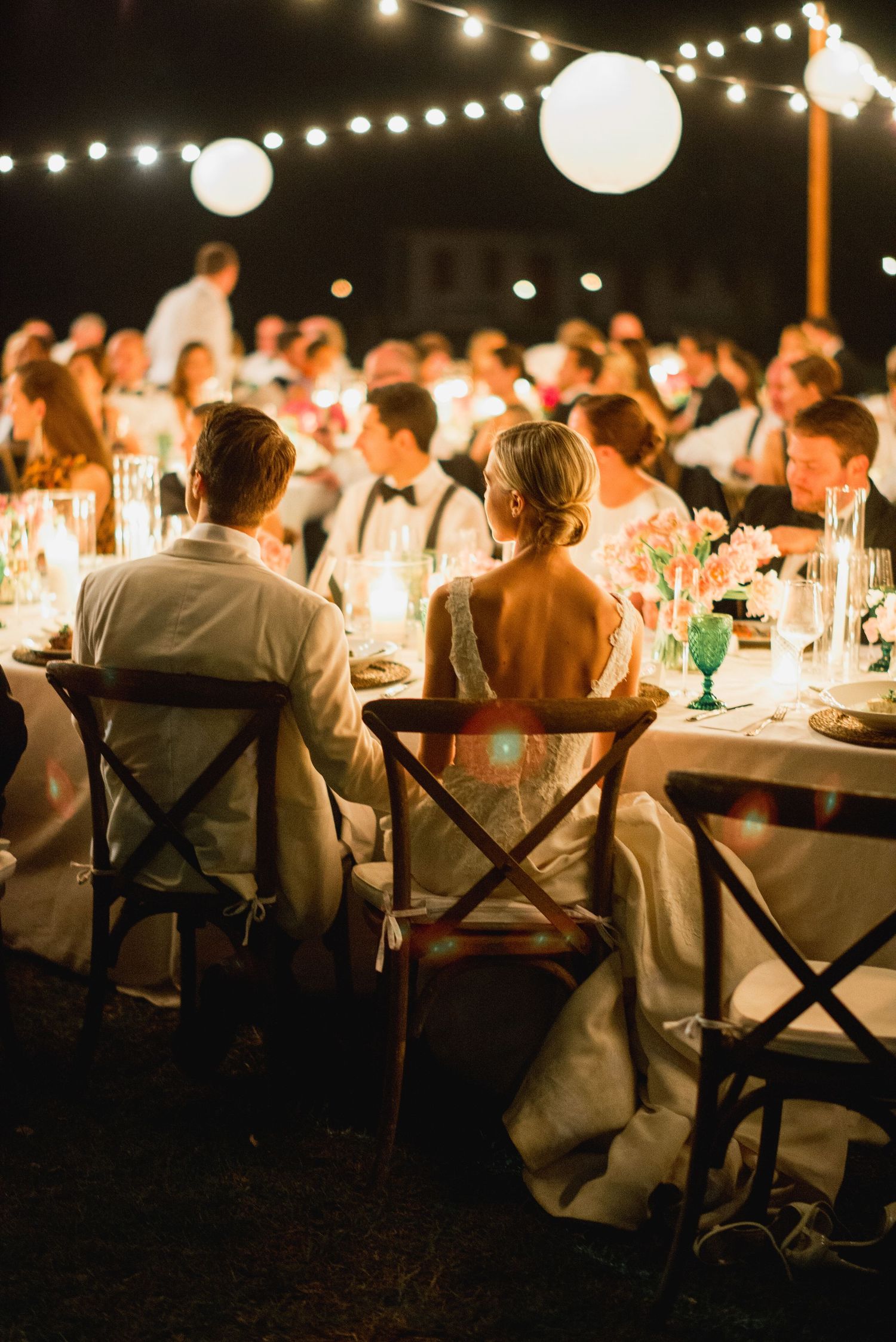 Wedding guests enjoy an intimate outdoor reception under string lights and paper lanterns at a rustic evening celebration.