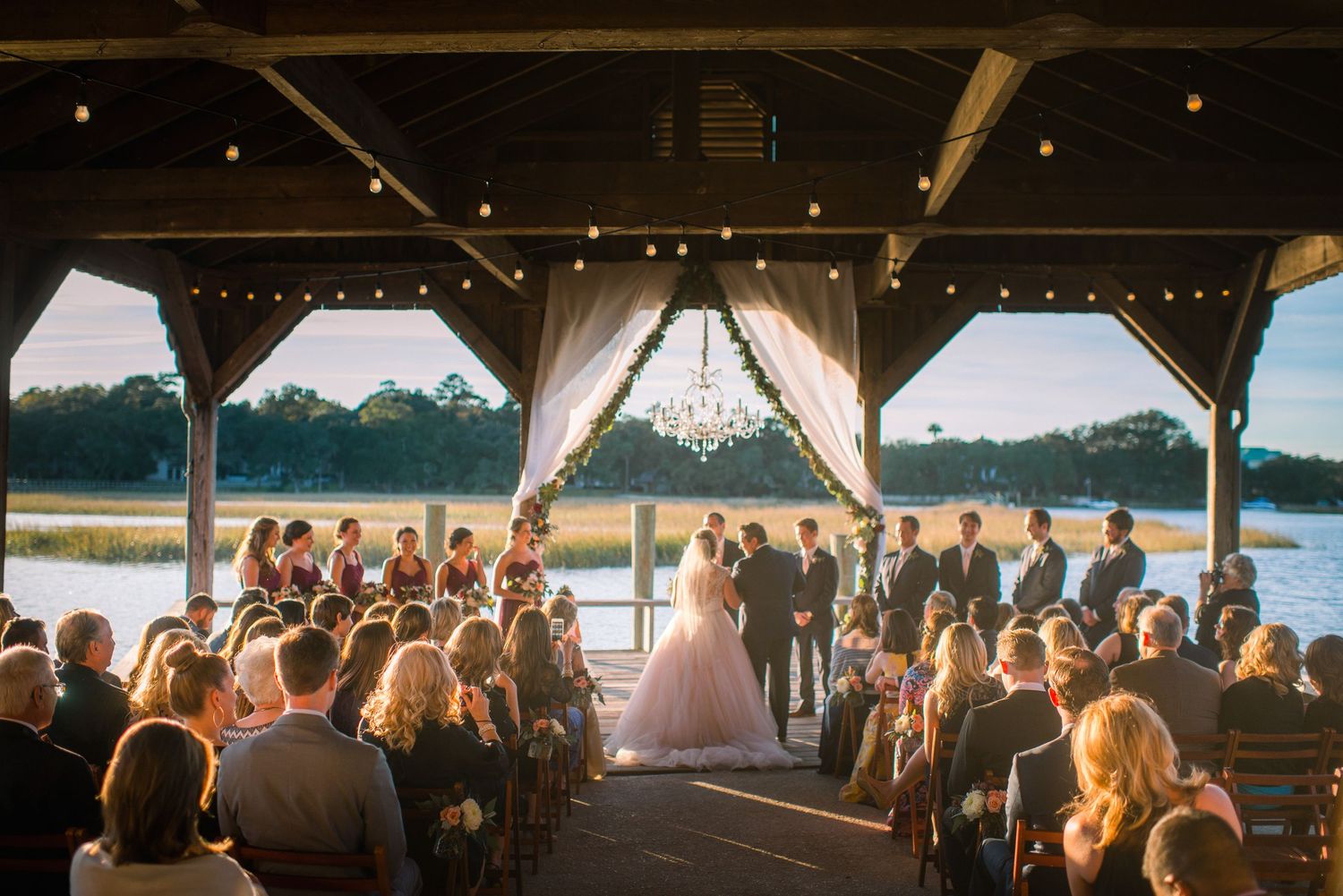 Wedding guests seated under a covered outdoor pavilion watching a lakeside ceremony at sunset.