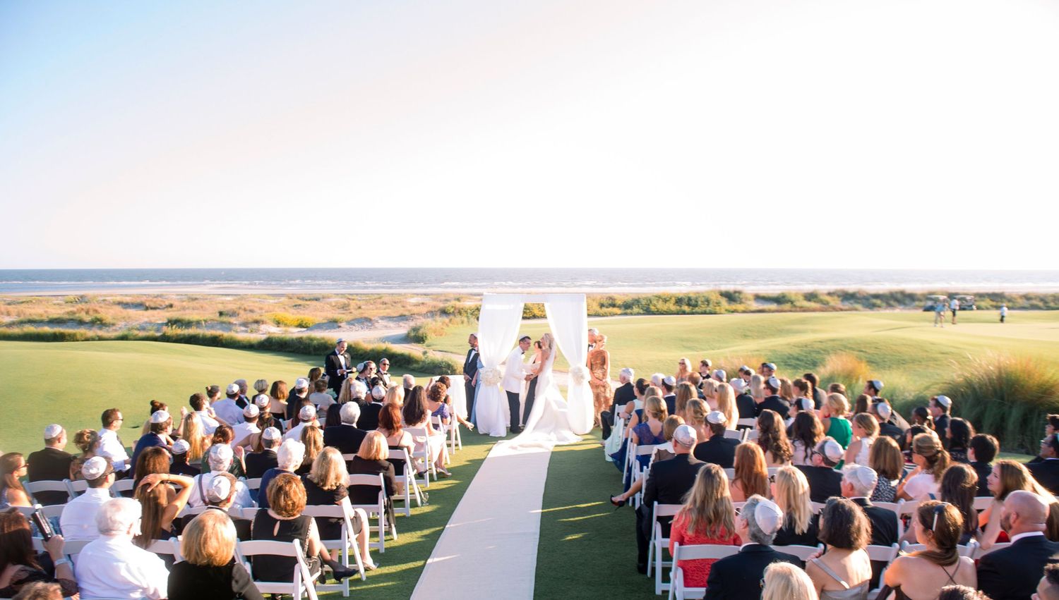 Panoramic view of an outdoor wedding ceremony setup with white chairs on a grassy hillside overlooking scenic farmland.