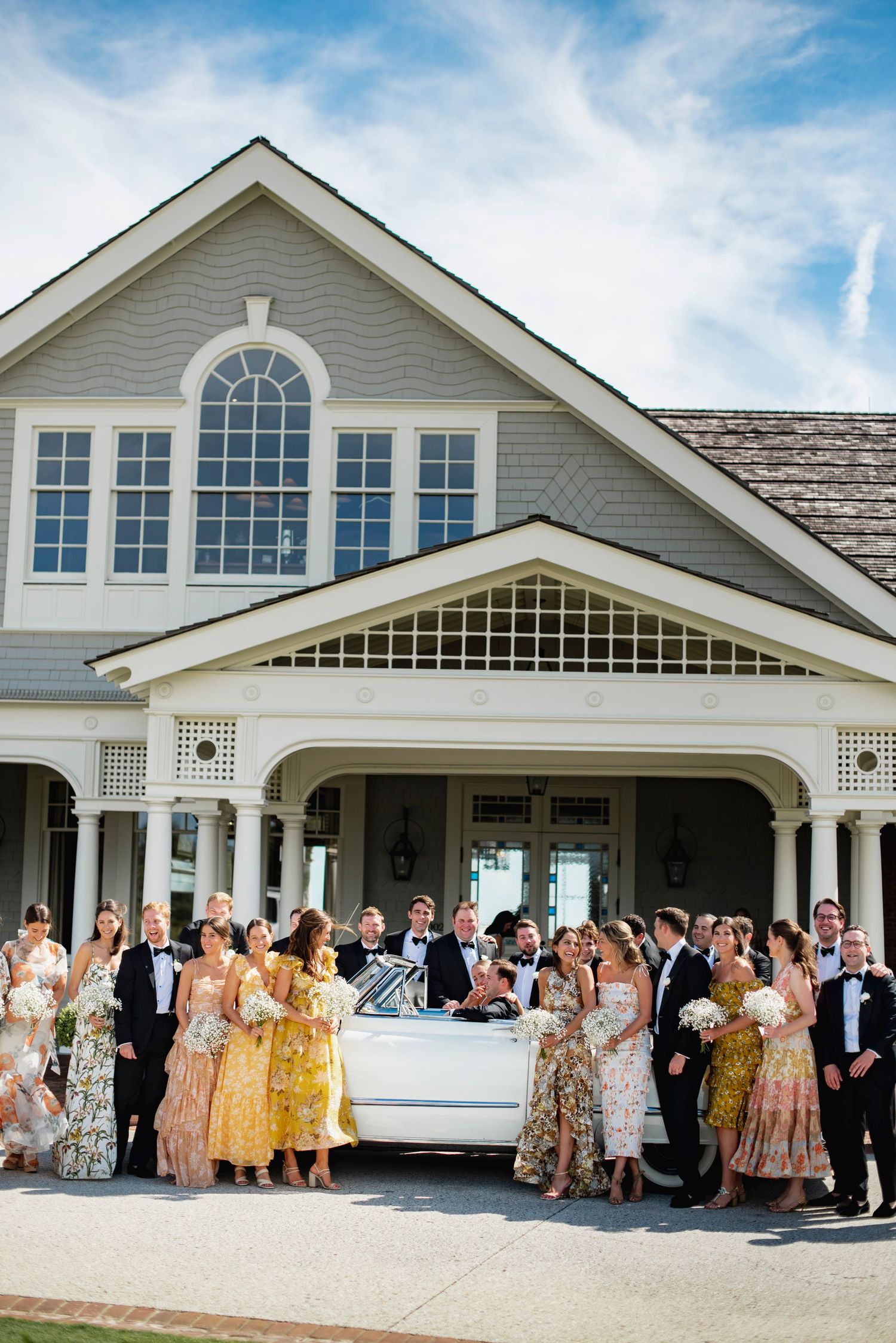 Wedding party in yellow and floral dresses gathered outside a white colonial-style venue with arched windows.