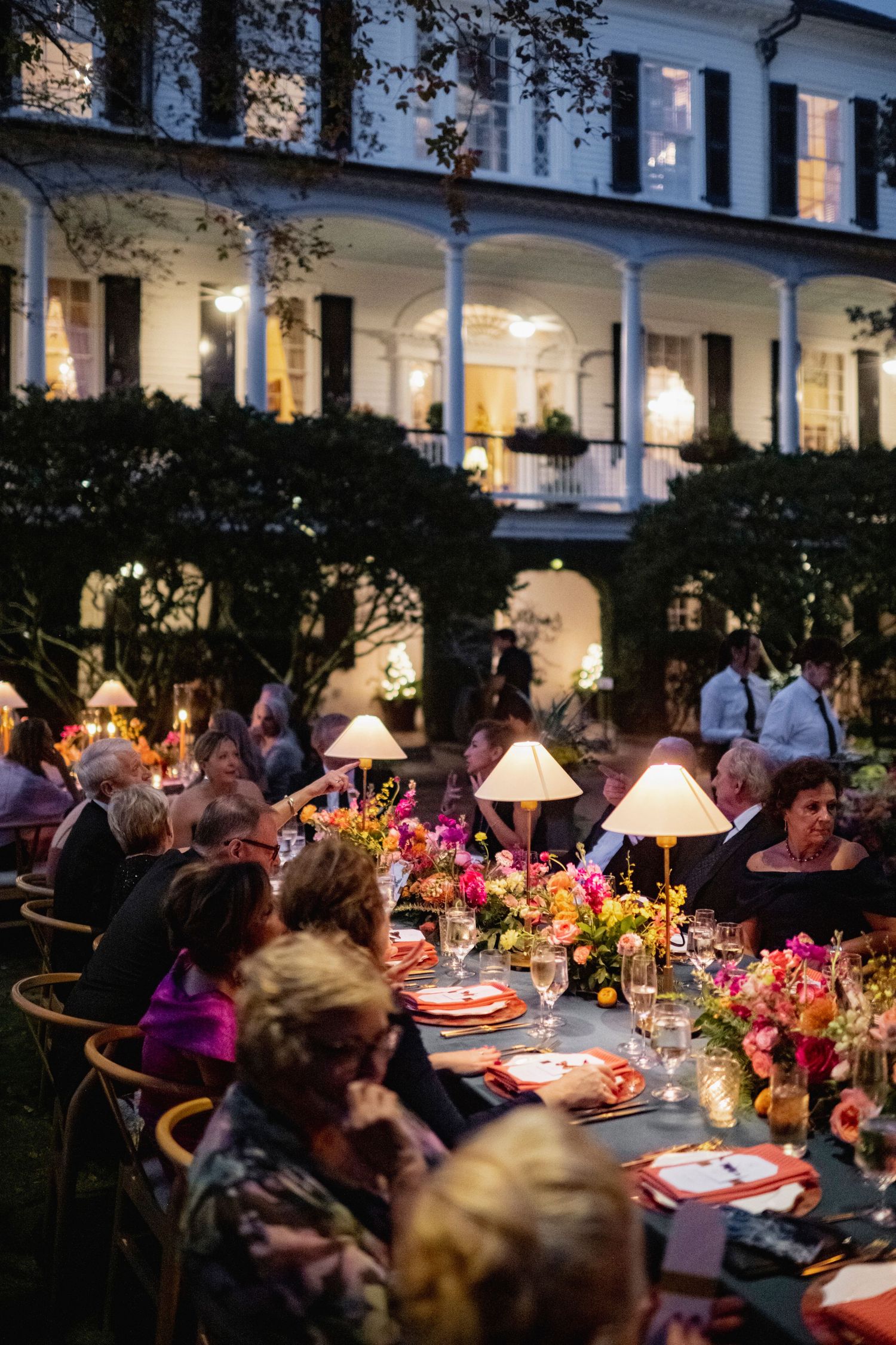Elegant outdoor evening dinner party with glowing lamps and floral centerpieces on long tables under a covered porch.