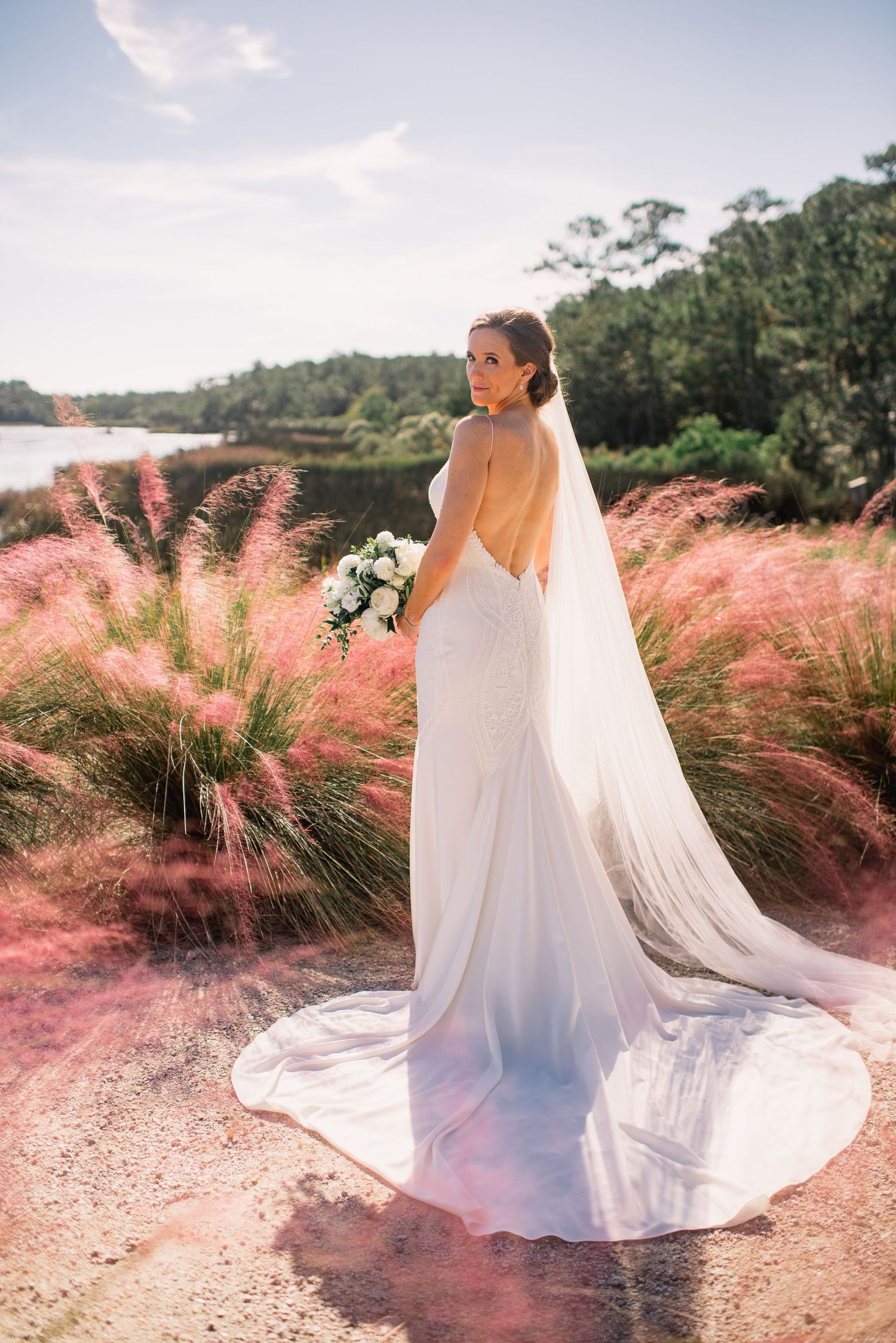 A flowing white wedding dress billows in the breeze against a backdrop of pink muhly grass at sunset.