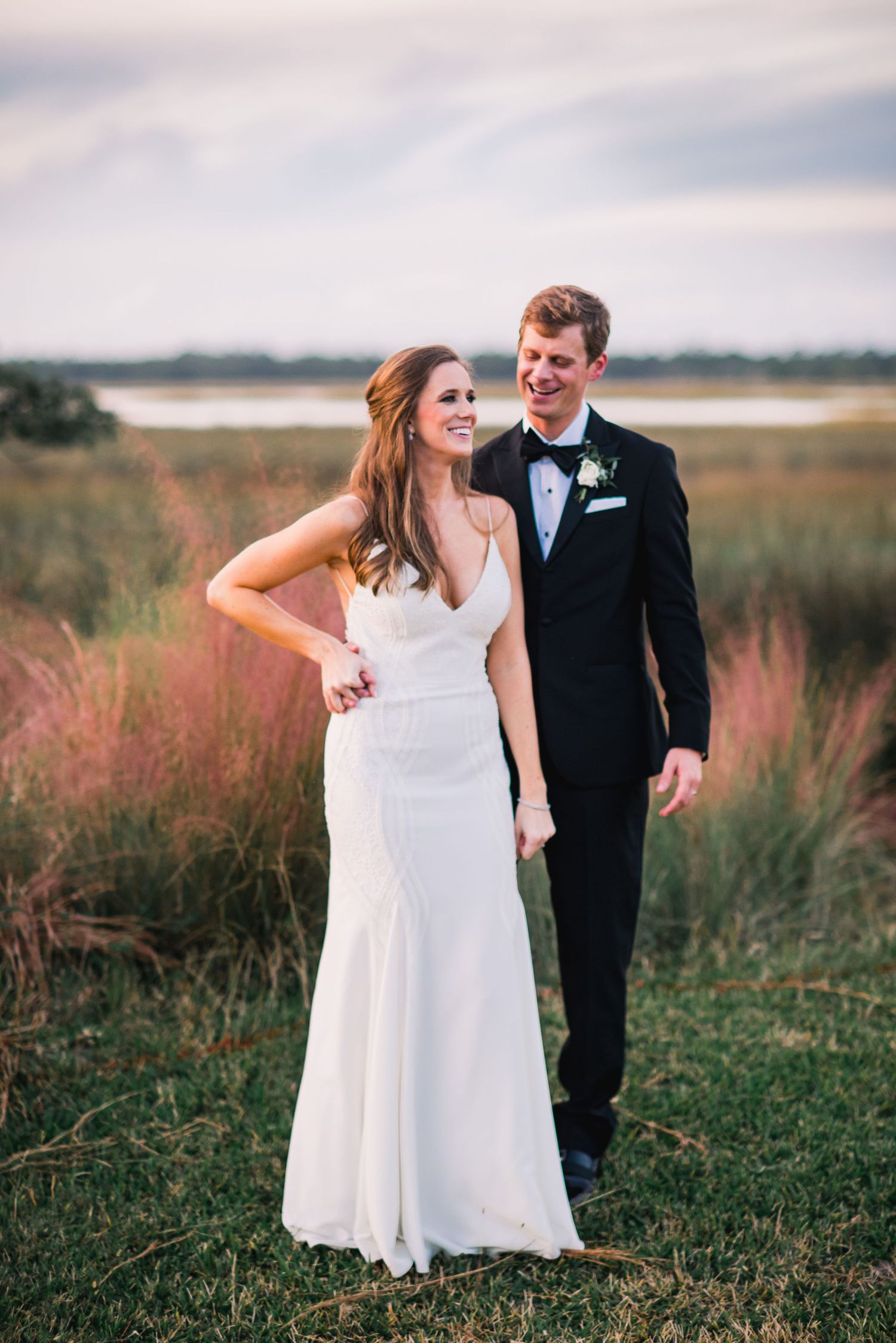 A couple in wedding attire shares a romantic moment in a grassy field with pink wildflowers at sunset.