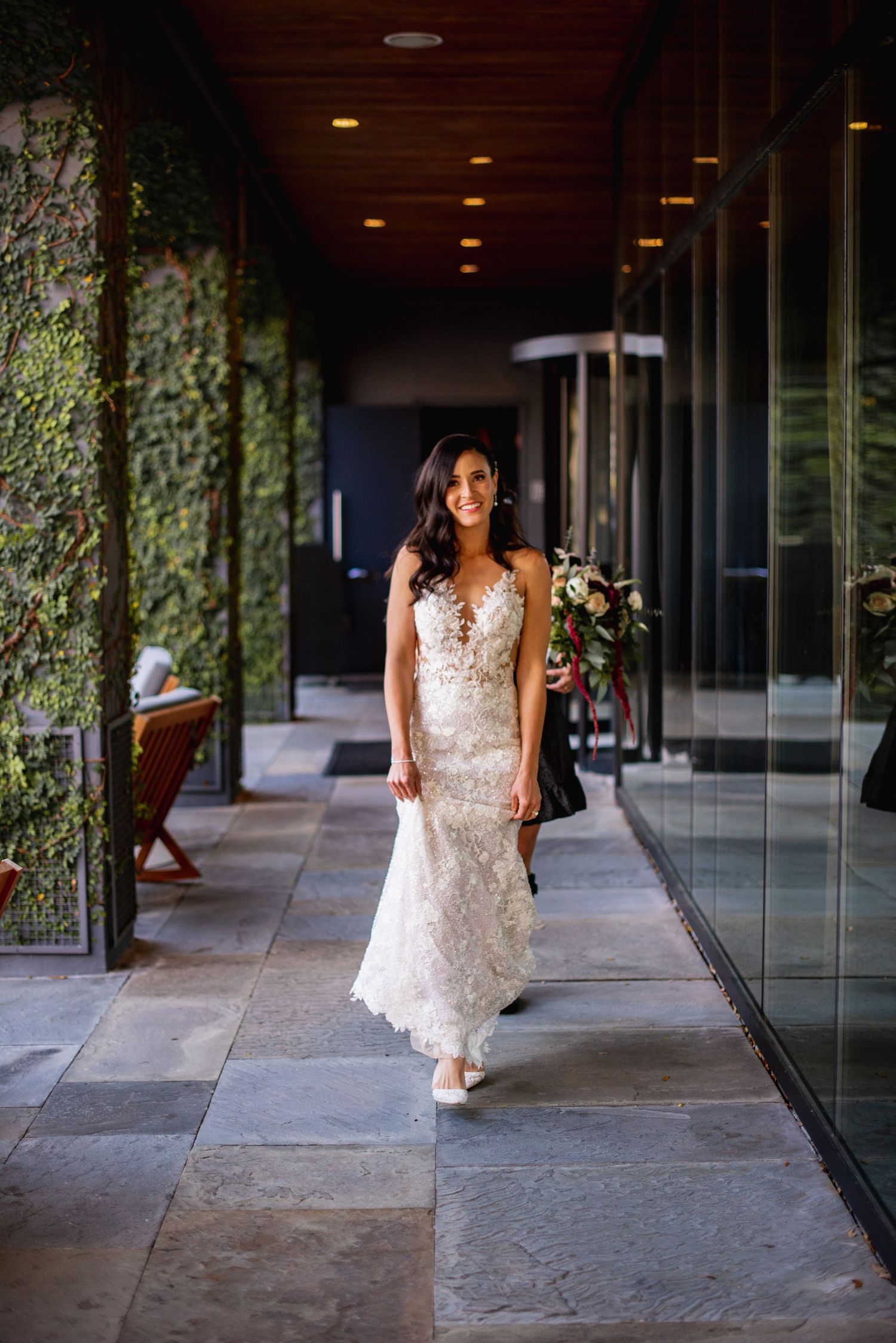 A person in a white lace wedding dress walks through a modern hallway with glass walls and ivy-covered columns.