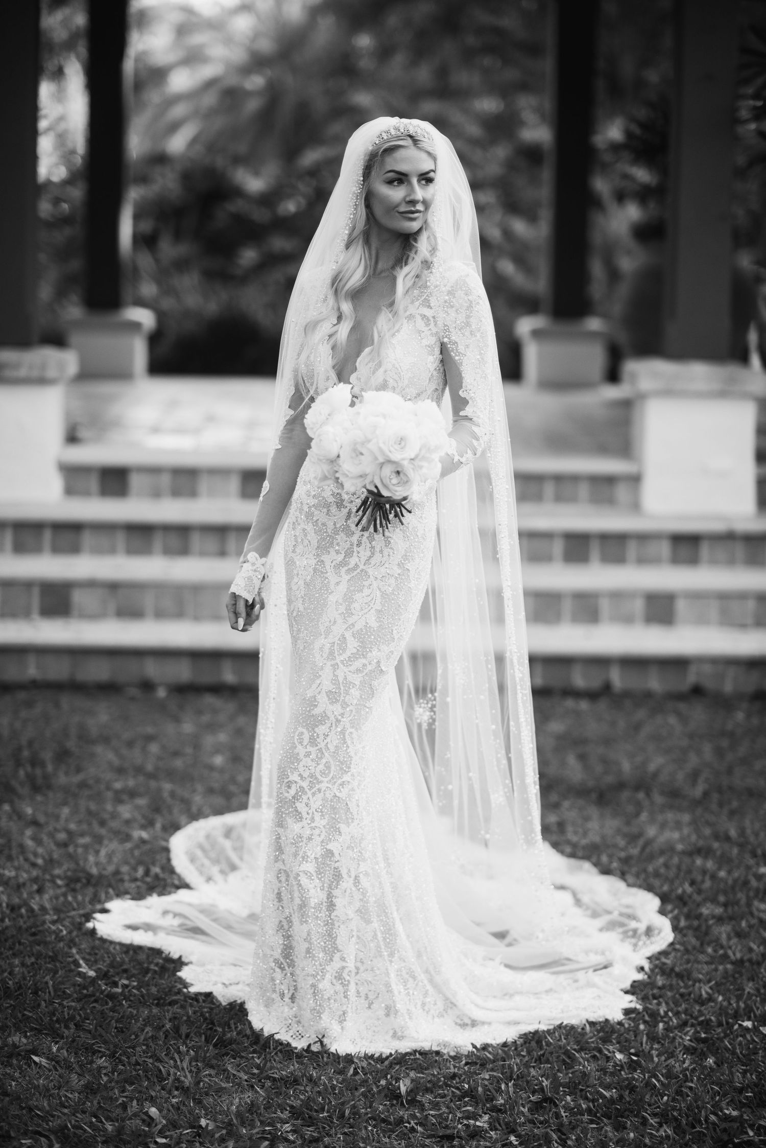 A bride in a white lace wedding gown and veil poses gracefully on grass for a series of black and white portraits.