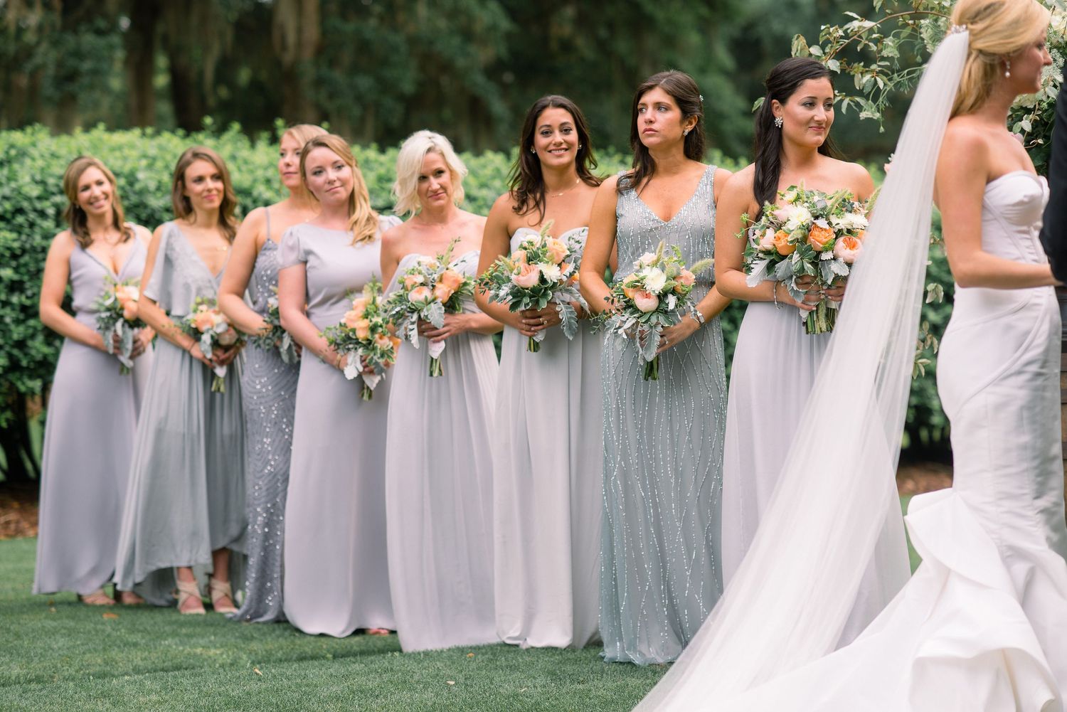 Wedding party in light gray bridesmaid dresses standing in line outdoors holding floral bouquets.