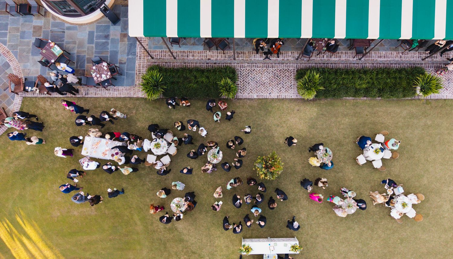 Aerial view of people gathered in small groups on a grass lawn next to a striped awning.