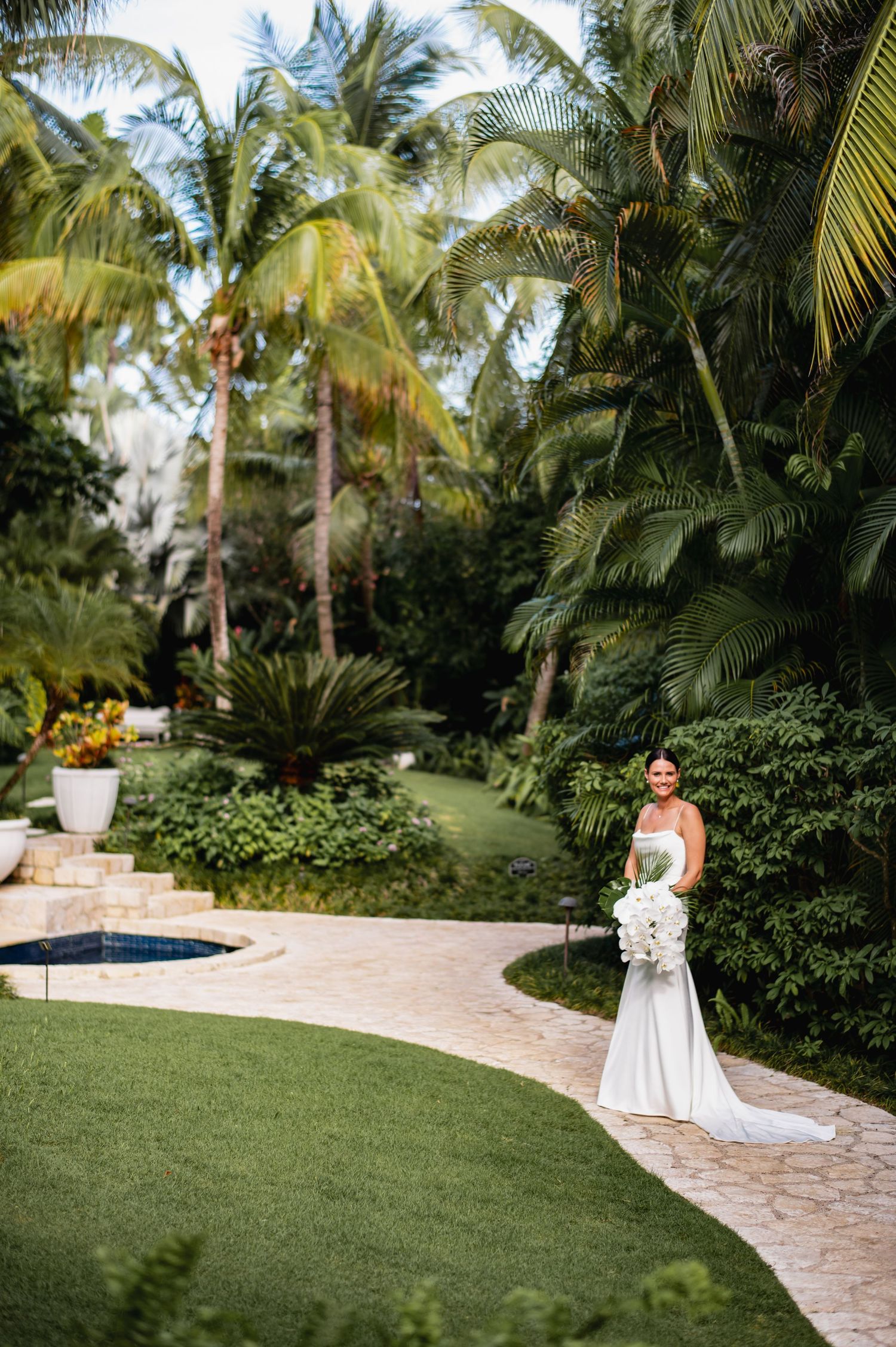 Lush tropical garden path lined with palm trees and greenery leads to a small pool area in a luxurious backyard setting.