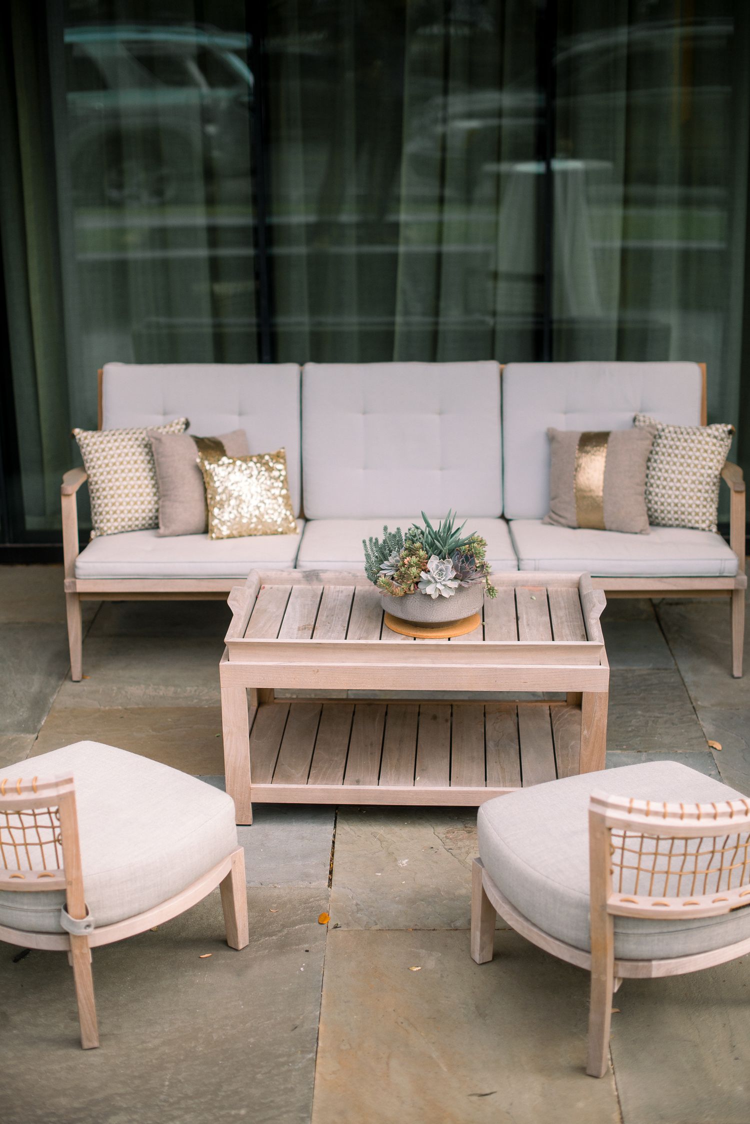 Modern outdoor patio furniture set featuring a light-colored wooden bench with plush cushions and decorative pillows.