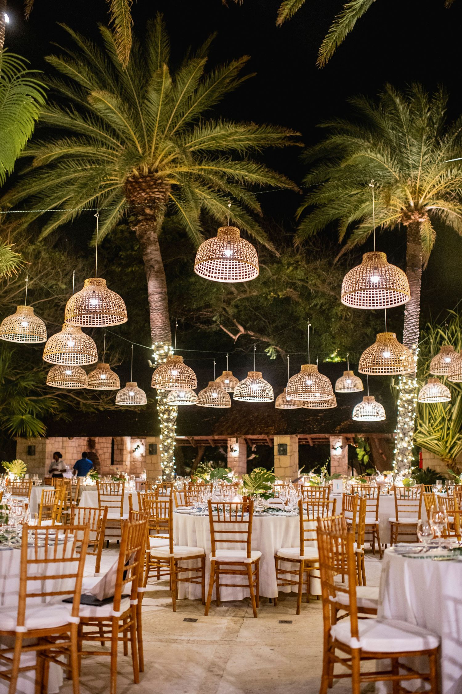 Elegant outdoor wedding reception setup with hanging wicker lanterns and palm trees illuminated at night.