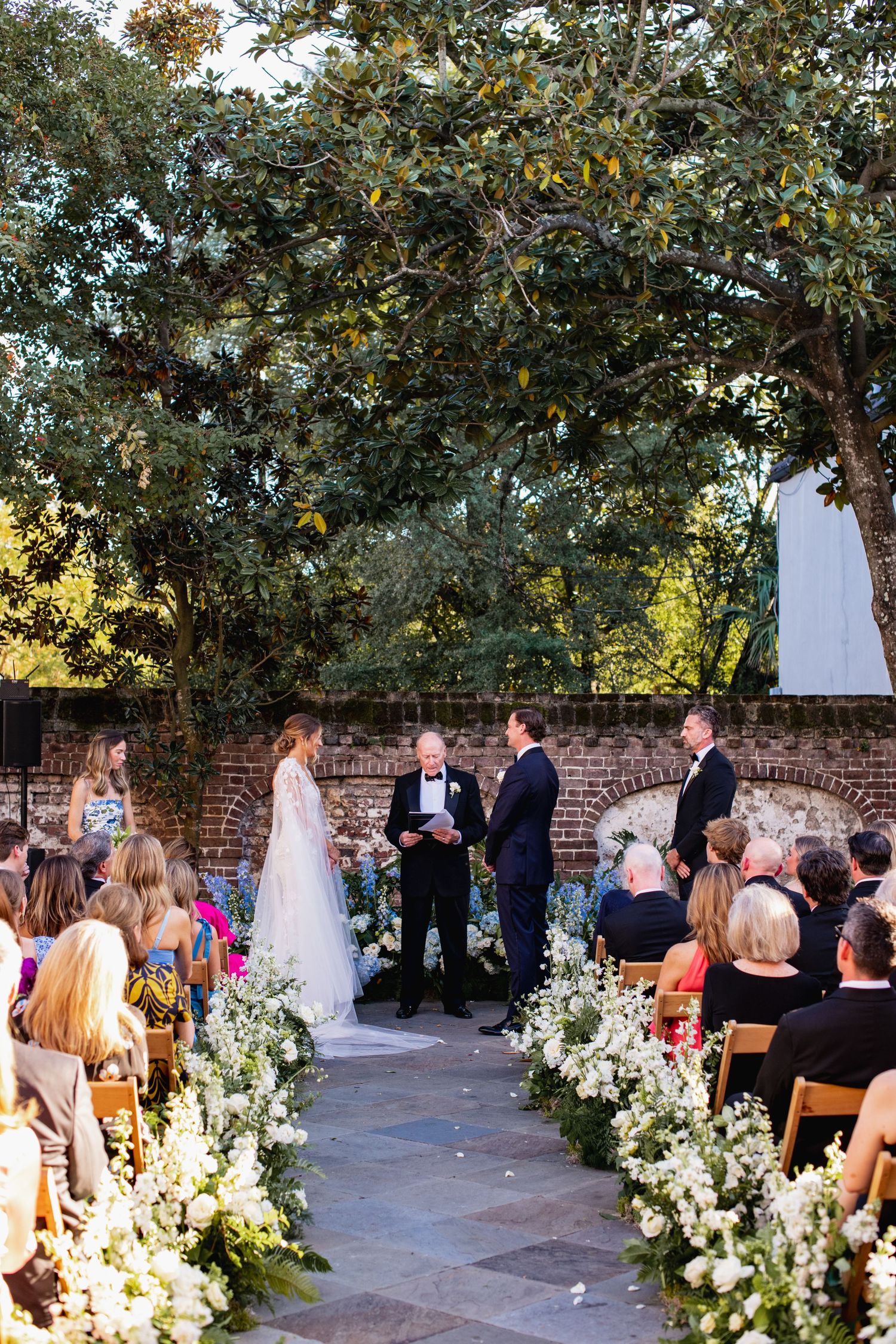 Outdoor wedding ceremony in a garden courtyard with brick walls, white floral arrangements, and guests seated on wooden chairs.