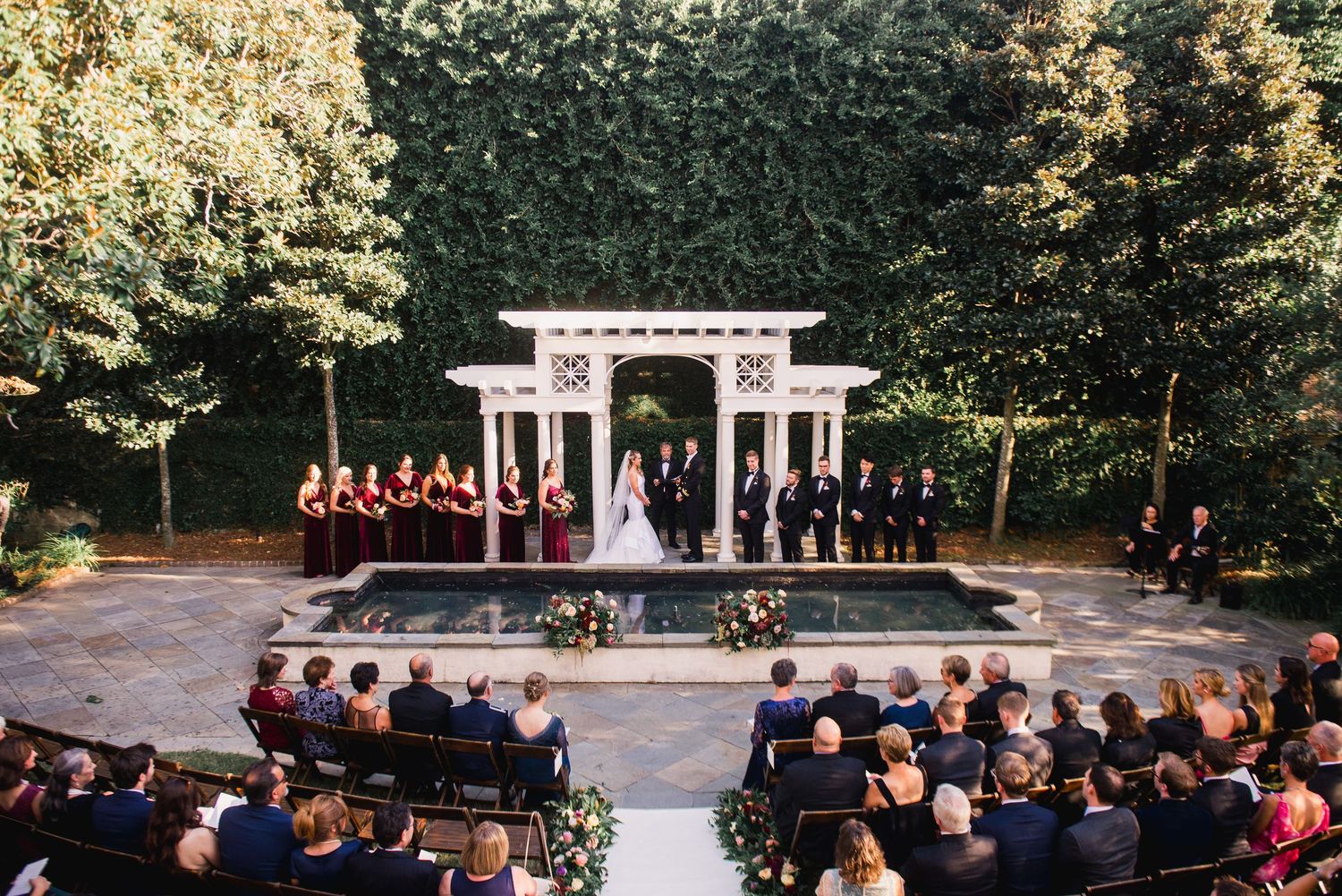 Wedding ceremony takes place under a white gazebo surrounded by lush greenery and guests seated outdoors.