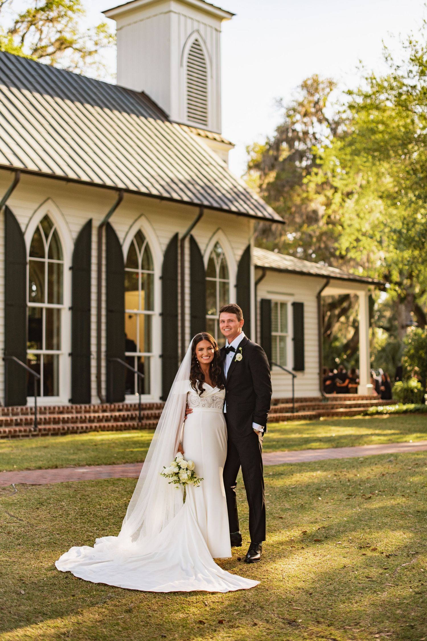 A wedding couple stands in front of a charming white chapel with Gothic windows and a metal roof on a sunny autumn day.
