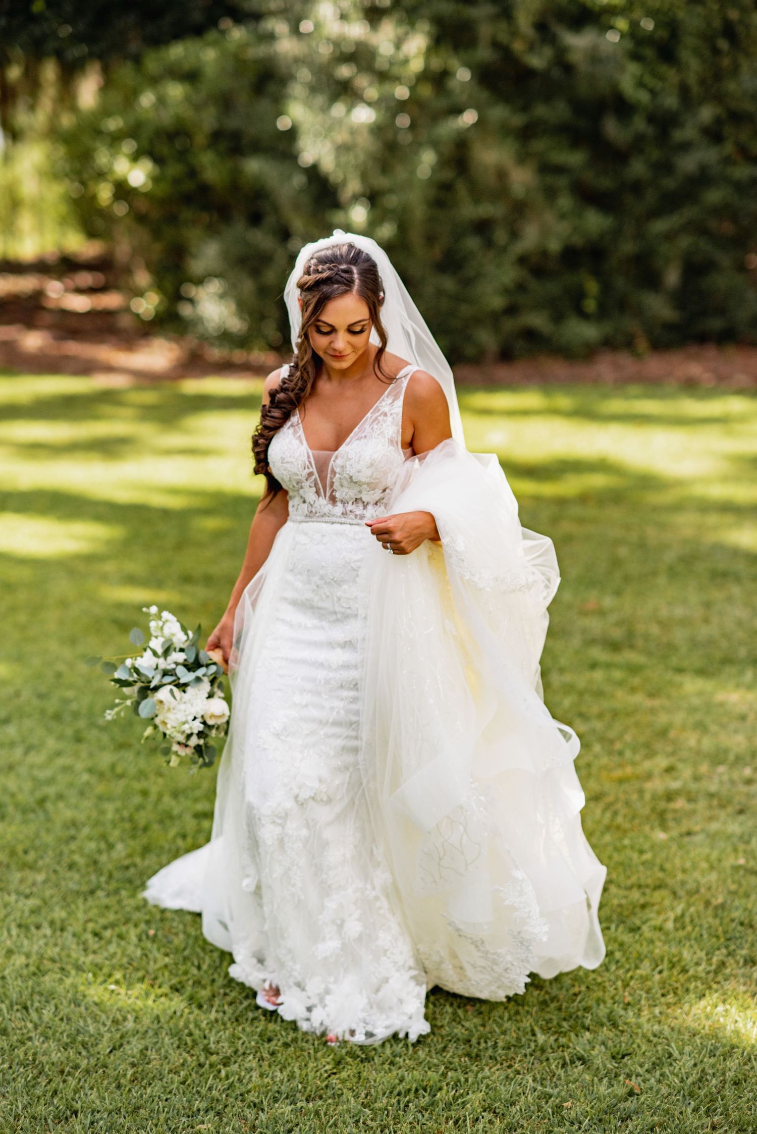 A bride in a white lace gown poses gracefully on lush green grass while holding a delicate bouquet.