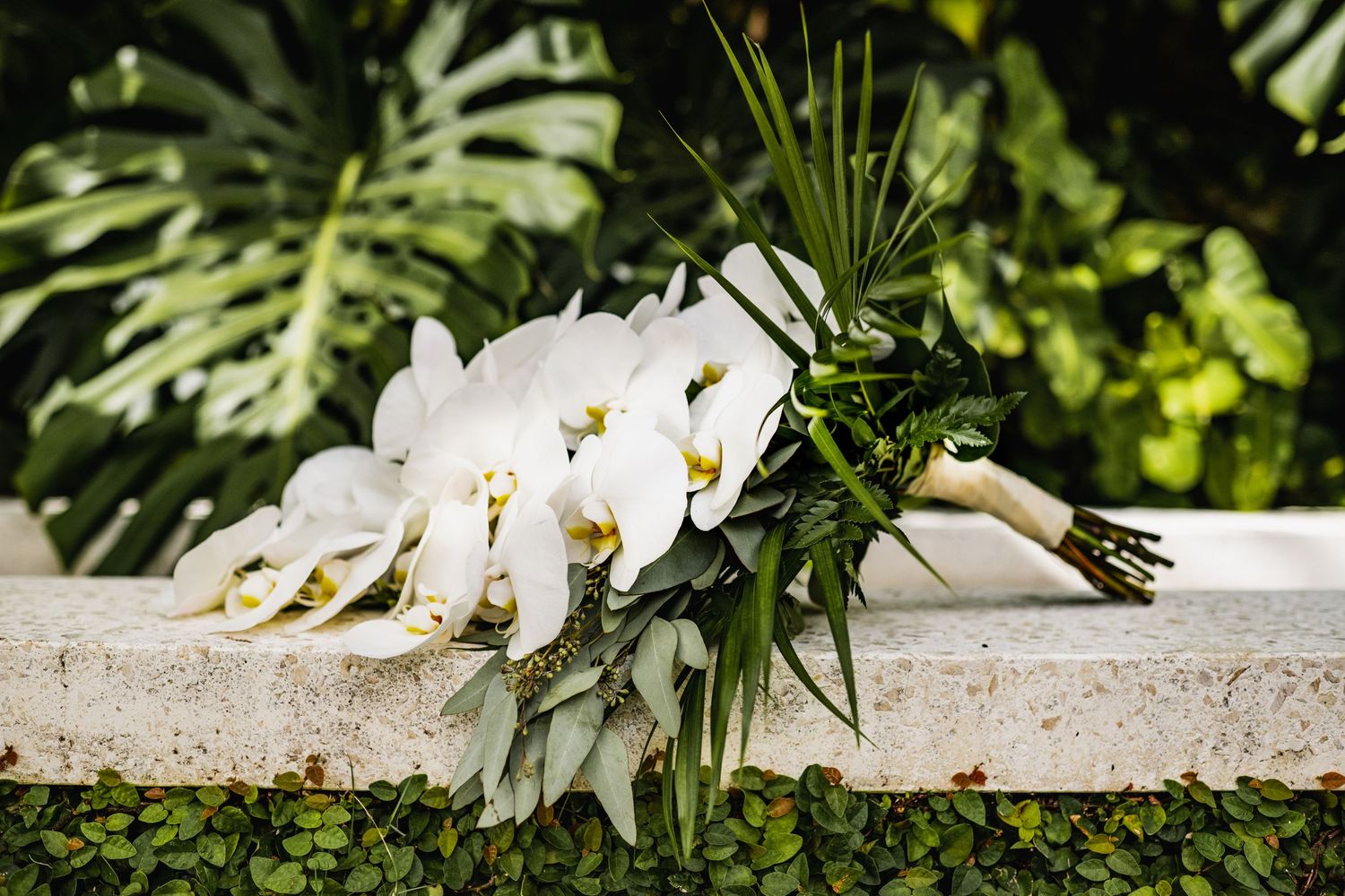 White orchid and tropical leaf wedding bouquet resting on a stone ledge with lush greenery in background.