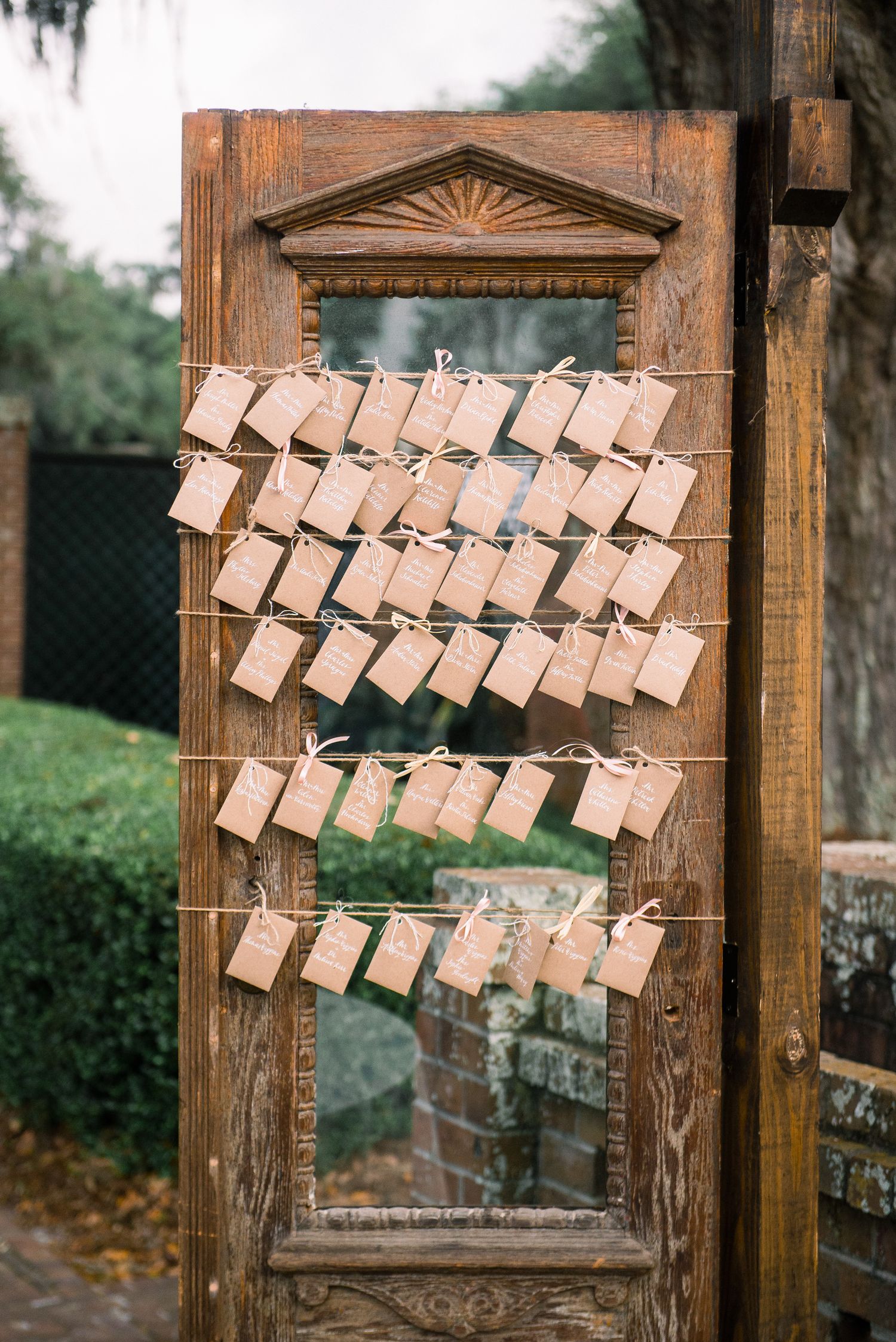 Brown paper escort cards with names hang from twine on a rustic wooden frame for an outdoor wedding display.