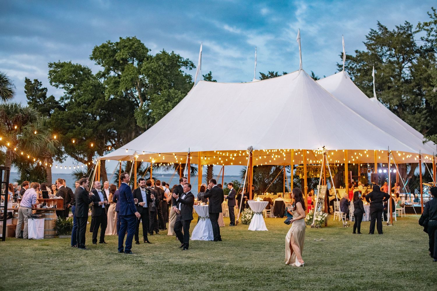 An elegant outdoor wedding reception under a white sailcloth tent with string lights and guests mingling on the lawn at dusk.
