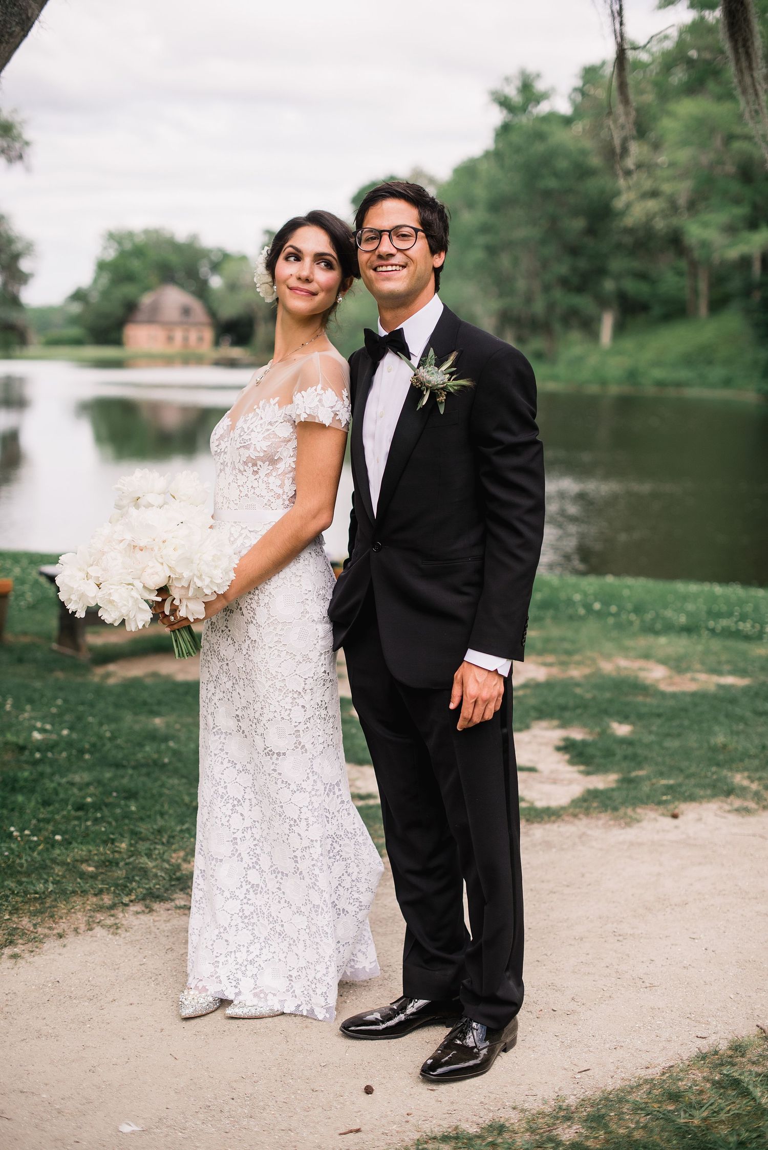 A romantic wedding couple poses by a serene lake, with the bride in a sparkling white gown and groom in classic black tuxedo.