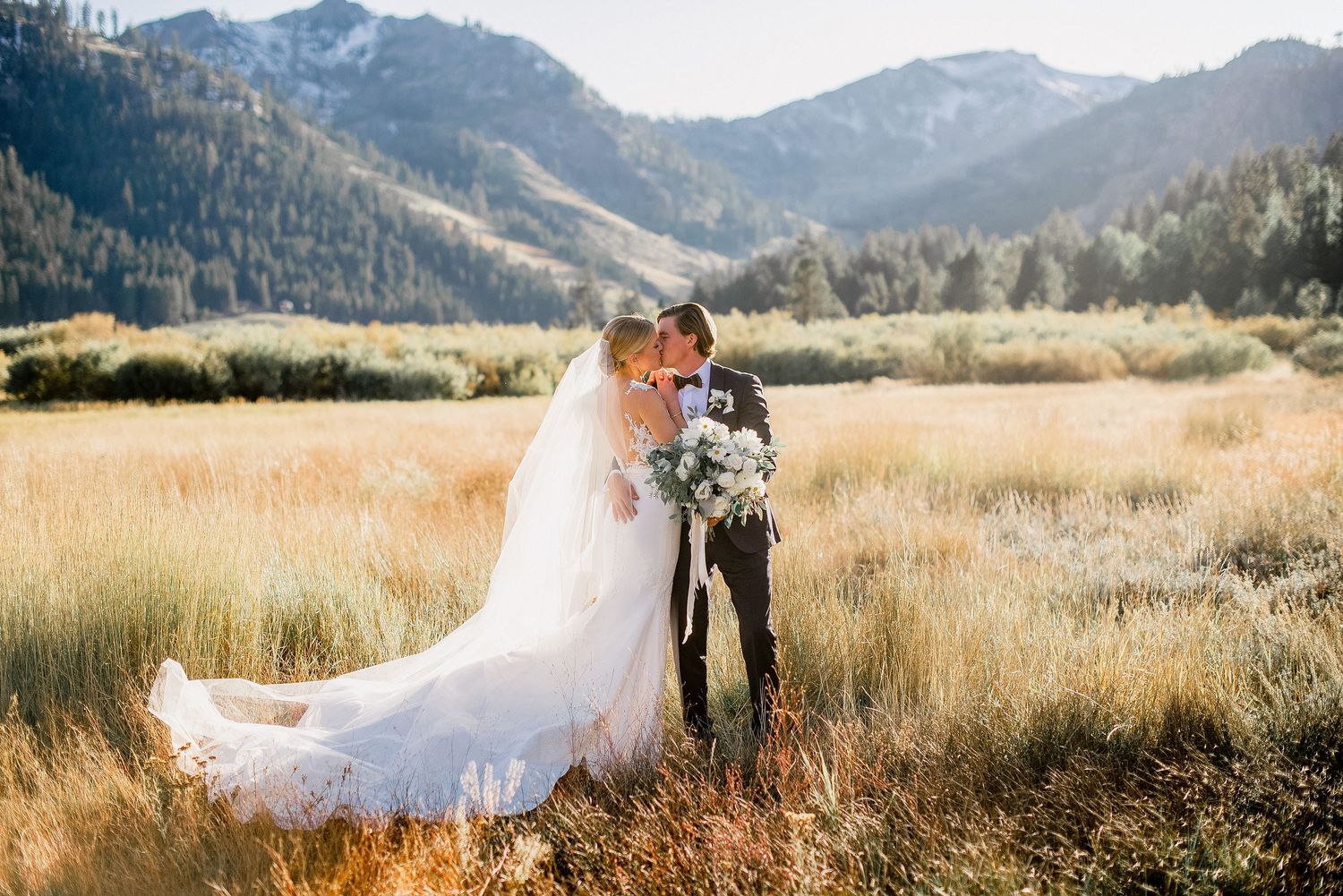 Couple embraces in golden mountain meadow with dramatic mountain peaks and evergreen forests in background during sunset.