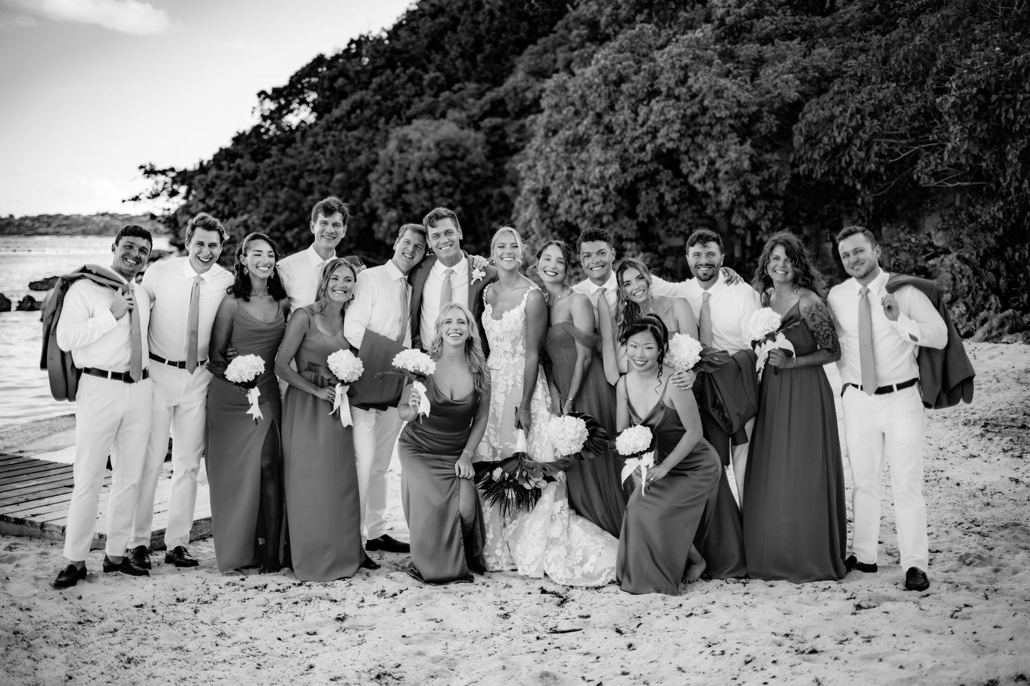Wedding party poses for formal group photo on beach in mix of formal attire.