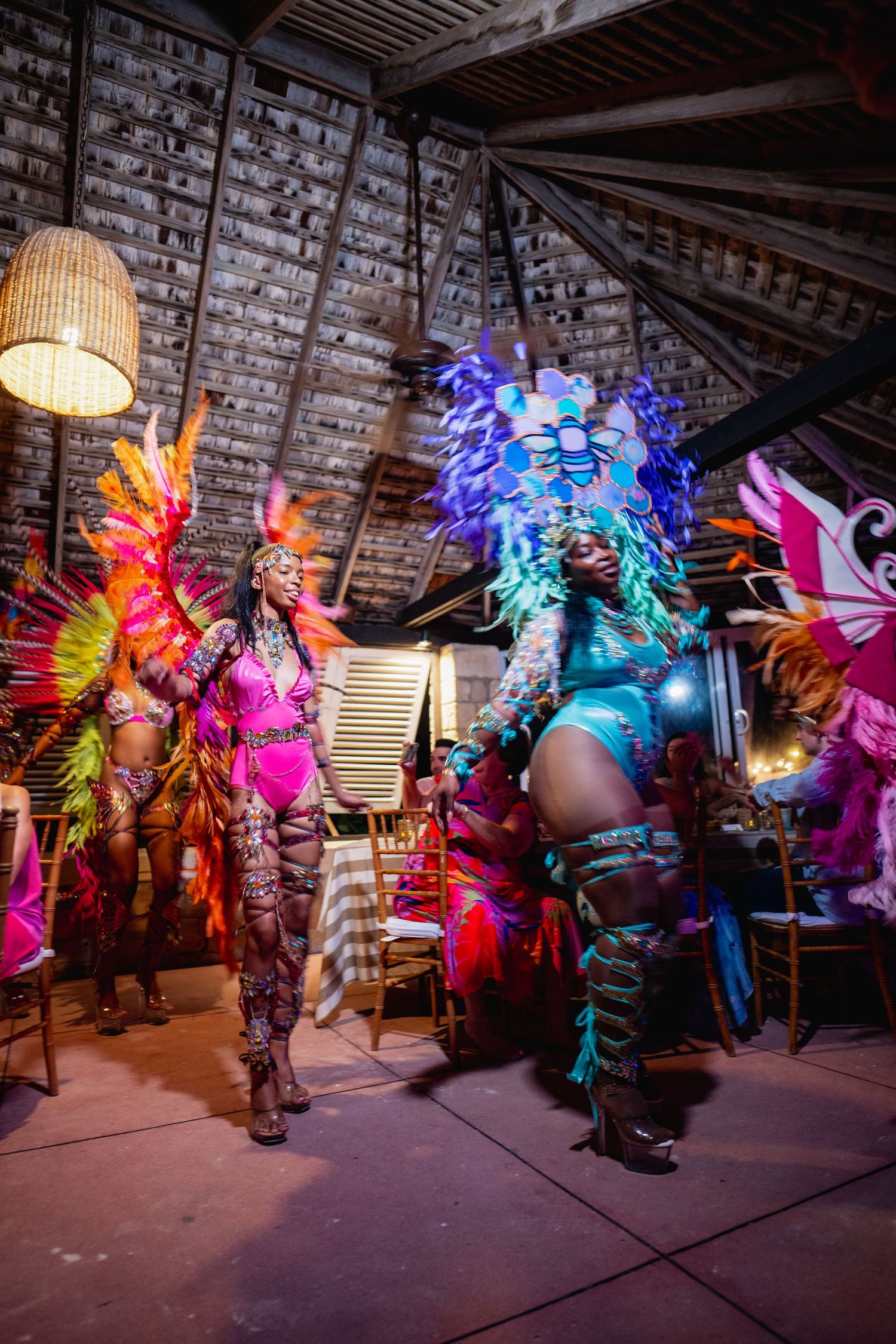 Colorful carnival performers in vibrant feathered costumes dancing under mood lighting at a tropical party.