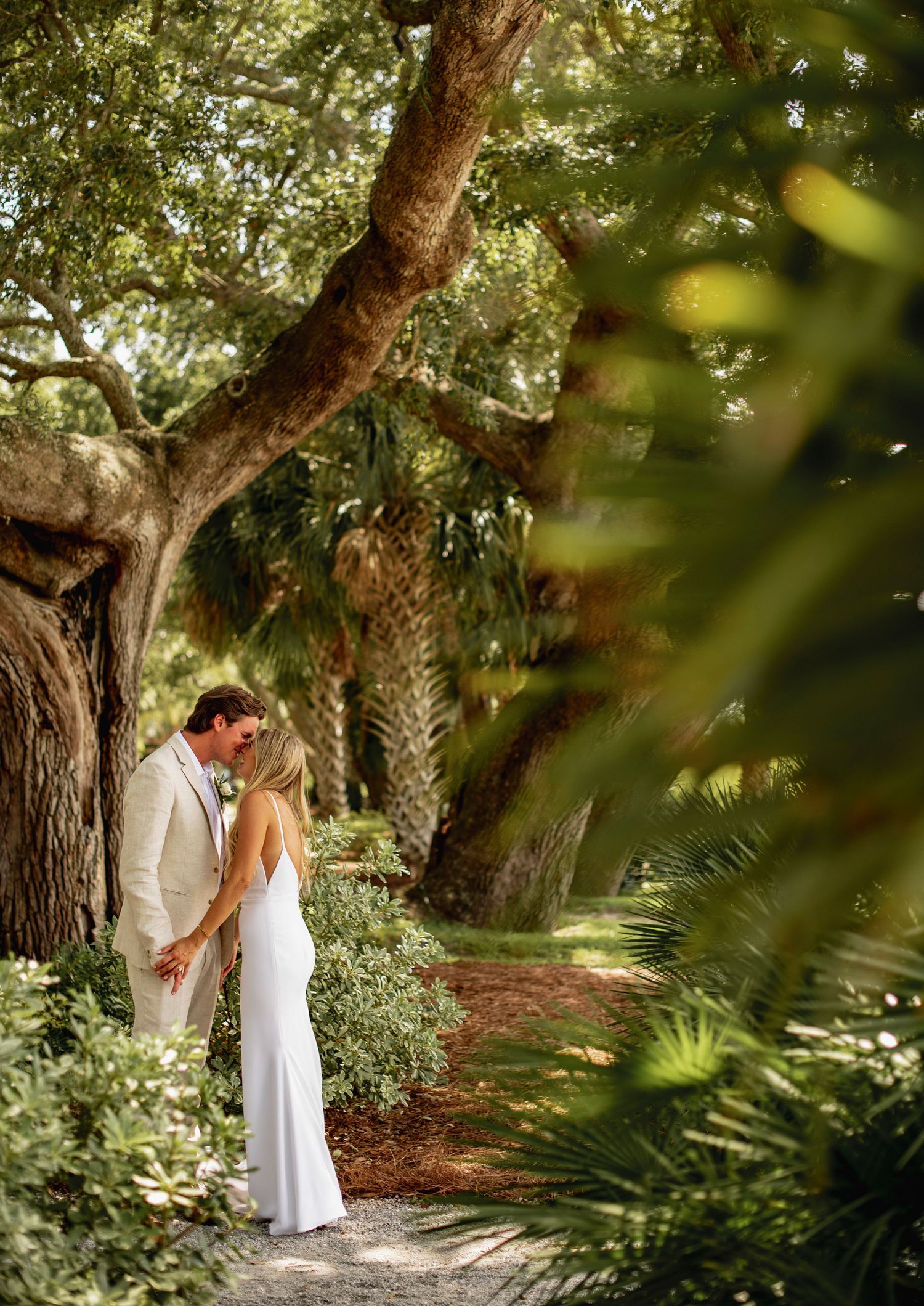 A couple shares a romantic moment on a garden path surrounded by lush trees and tropical foliage.