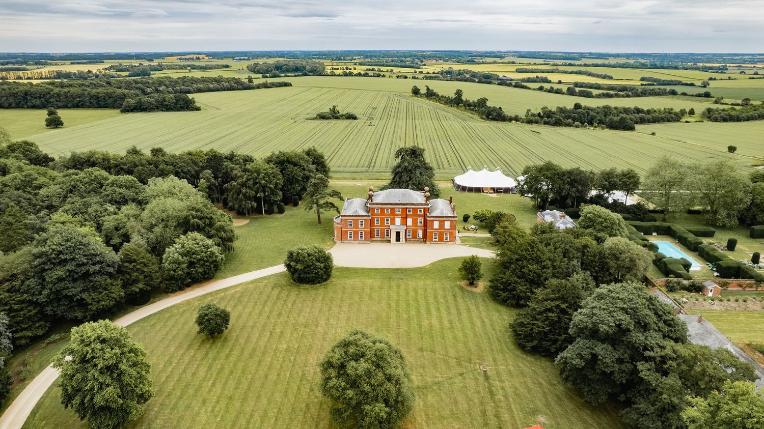Aerial panoramic view of lush green farmland and countryside estates with scattered trees and buildings under blue sky.