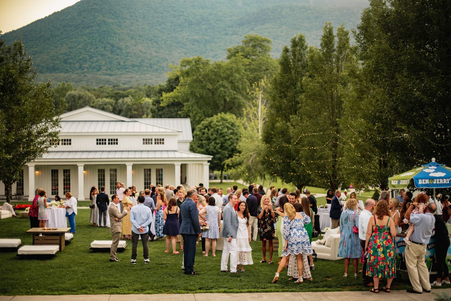 Wedding guests gather on a lush green lawn outside white buildings during an outdoor summer celebration.