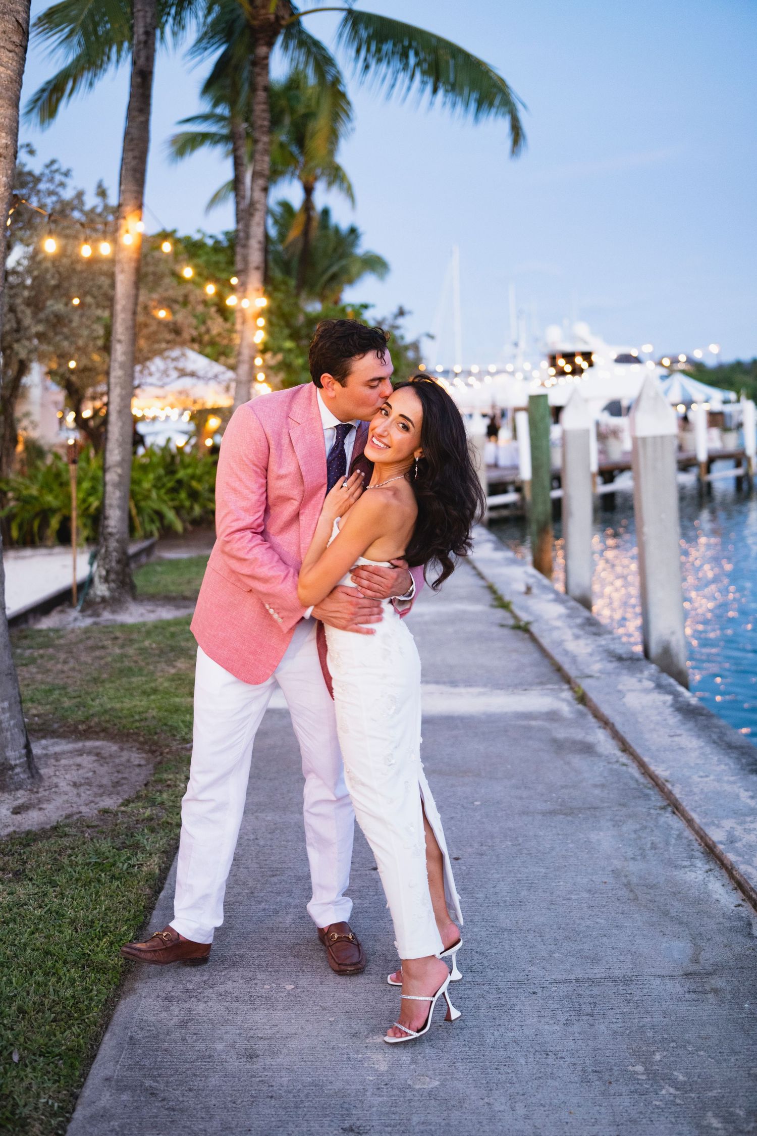 Romantic couple shares tender moments along a marina dock at sunset with palm trees and boats in the background.