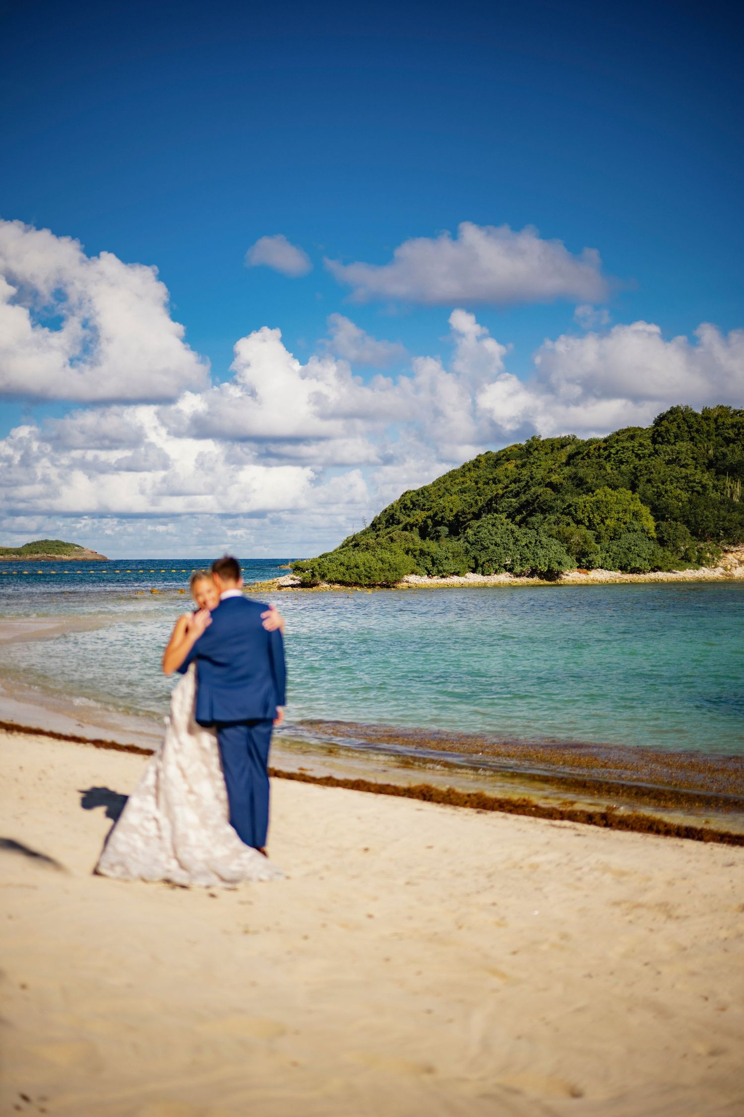 A couple in wedding attire shares a romantic moment on a tropical beach with turquoise waters and lush islands in view.