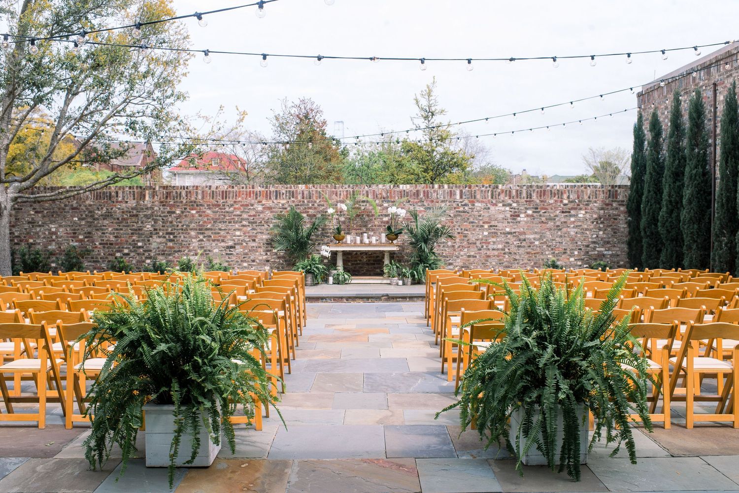 Outdoor wedding ceremony setup with wooden chairs and lush fern arrangements against a rustic stone wall.