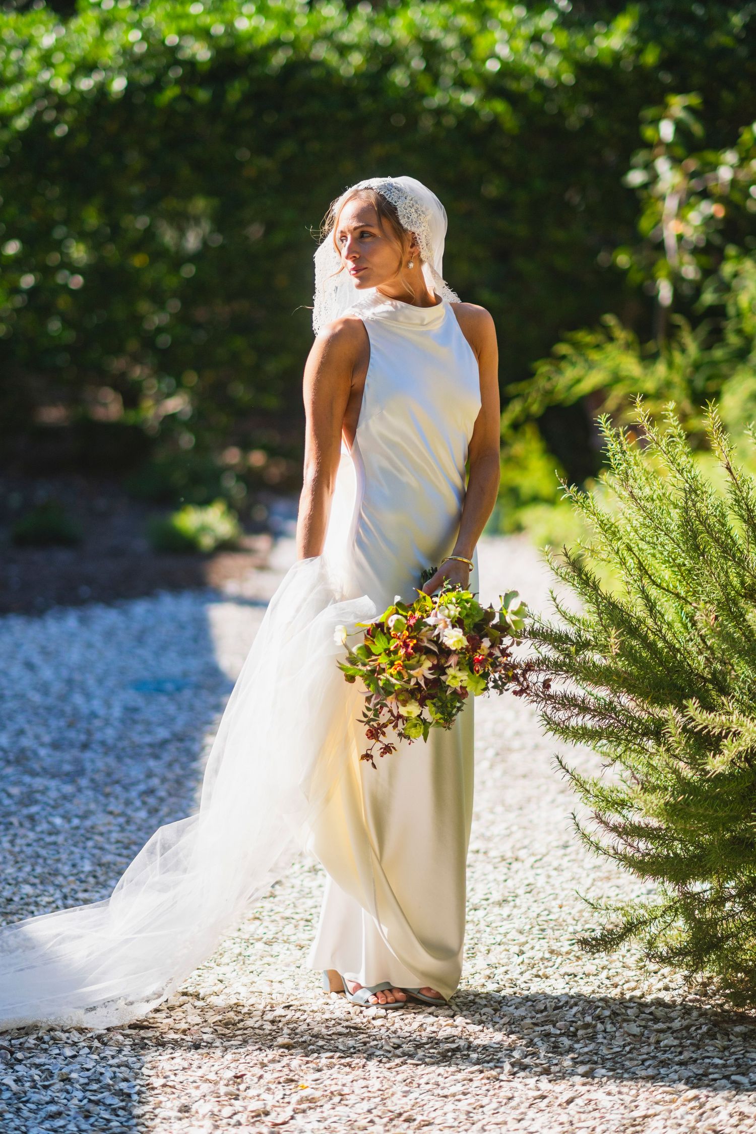 A bride in a white dress sits gracefully on a stone path surrounded by lush greenery and flowers in a garden setting.