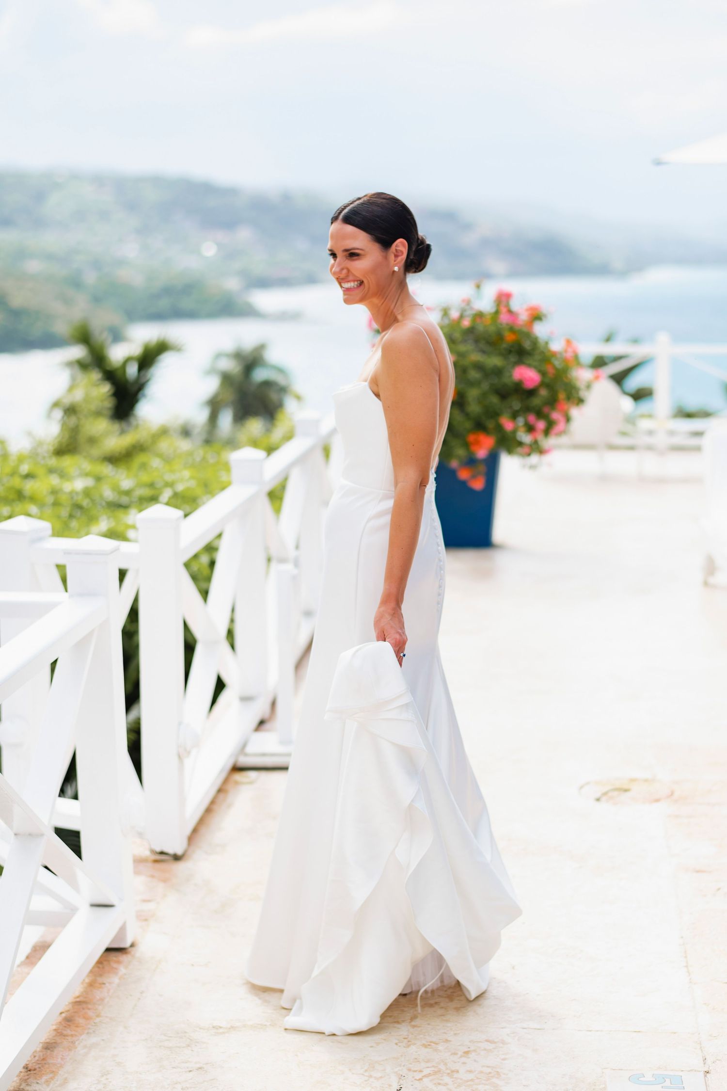 Bride in flowing white dress walking along tropical white balcony with ocean view.