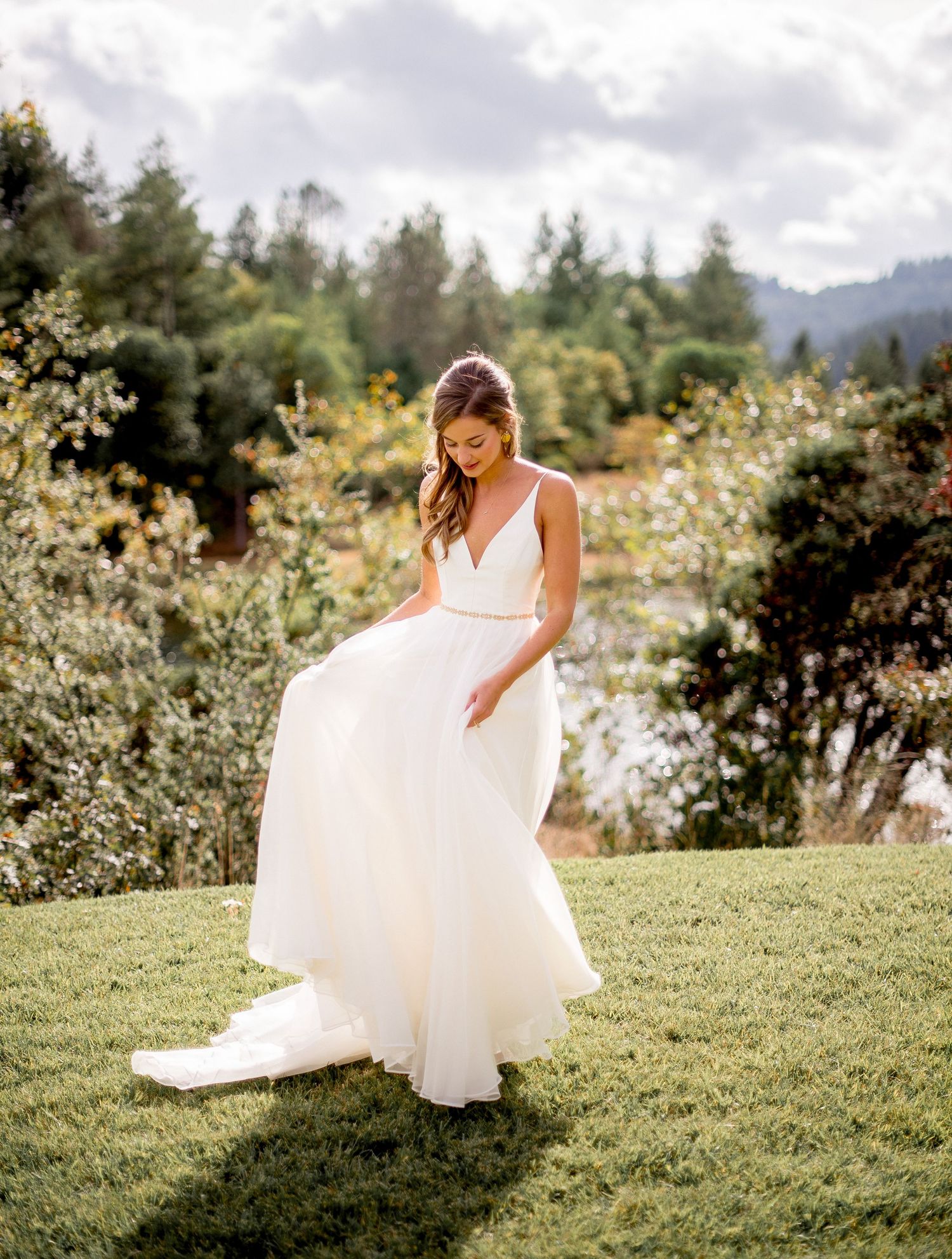 A bride in a flowing white dress twirls gracefully on a sunlit grassy hillside surrounded by trees and mountains.