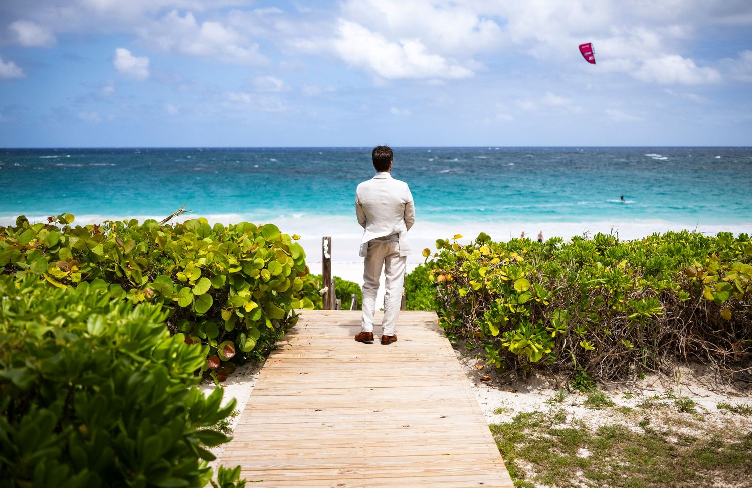 Person in white clothing walks on wooden boardwalk towards turquoise ocean with red kite in the sky.