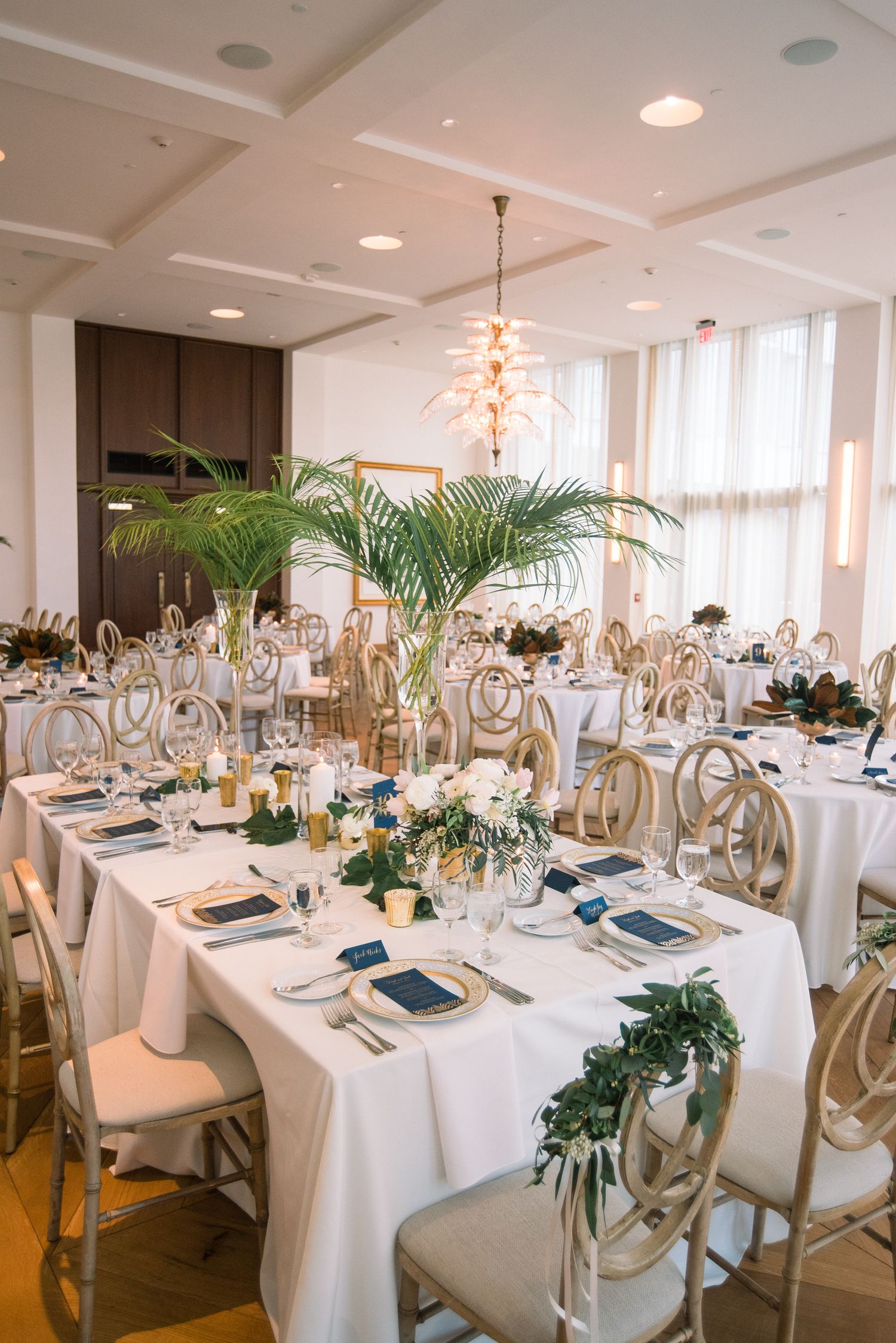 Elegant wedding reception tables set with white linens, gold chairs, and tropical greenery centerpieces in a bright venue.