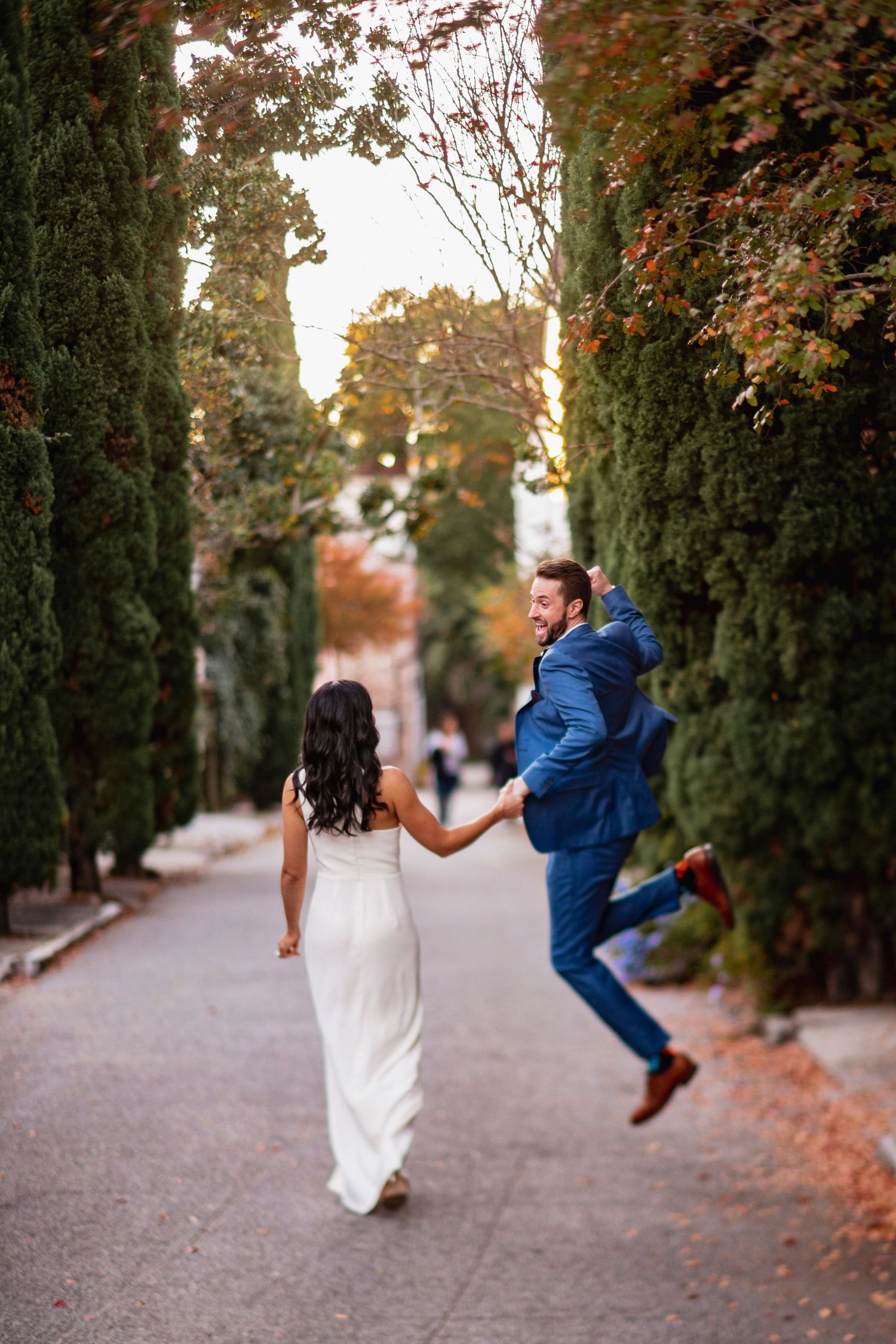 Playful wedding couple dancing and jumping in tree-lined path during autumn.