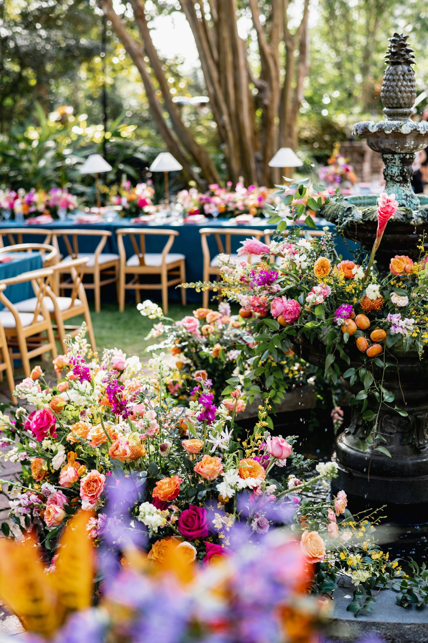 Vibrant floral arrangements with pink, orange and purple blooms decorate outdoor wedding tables under trees.