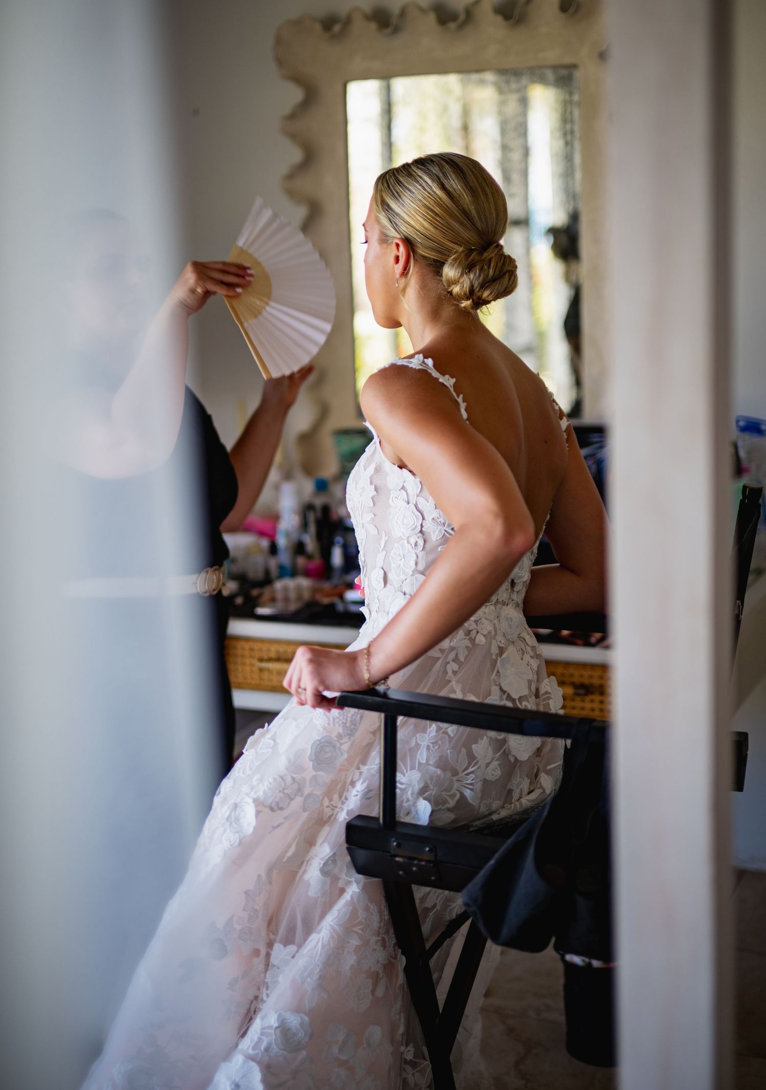 A bride in a white lace dress gets ready in a sunlit room with a handheld fan.