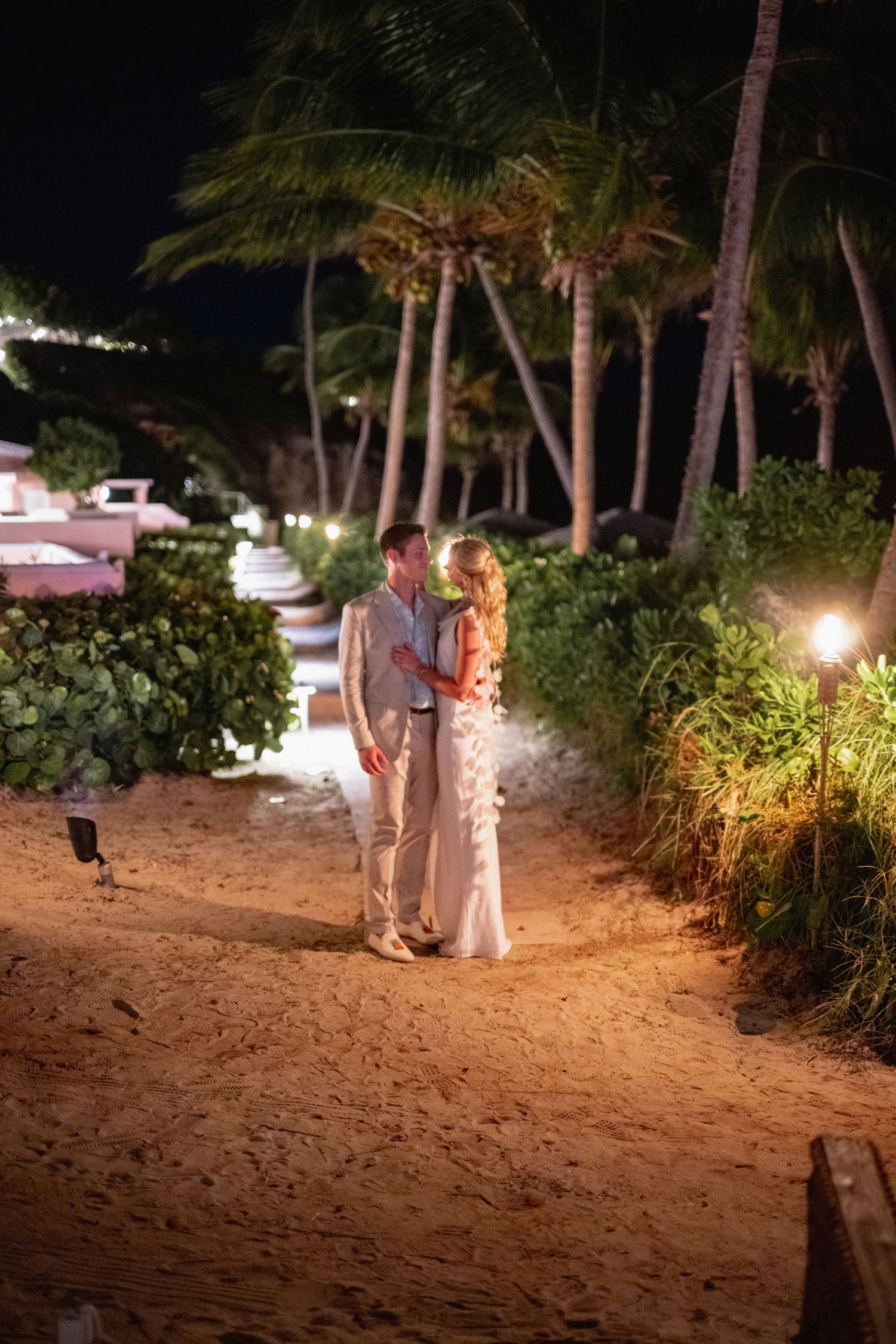 A couple walks along a sandy beach path at night with palm trees and landscape lighting creating a romantic ambiance.