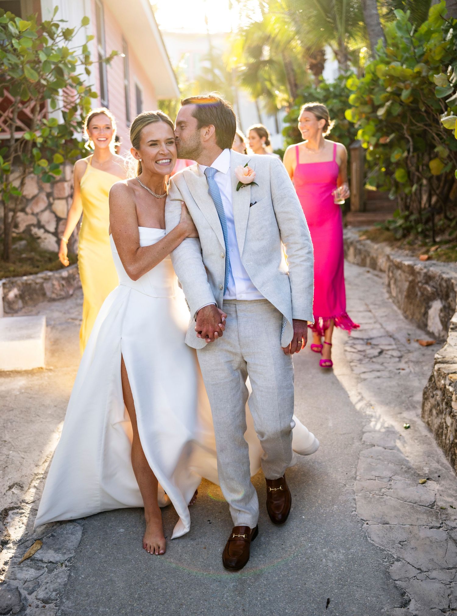 A couple in wedding attire walking down a charming cobblestone path with guests in bright summer clothing.