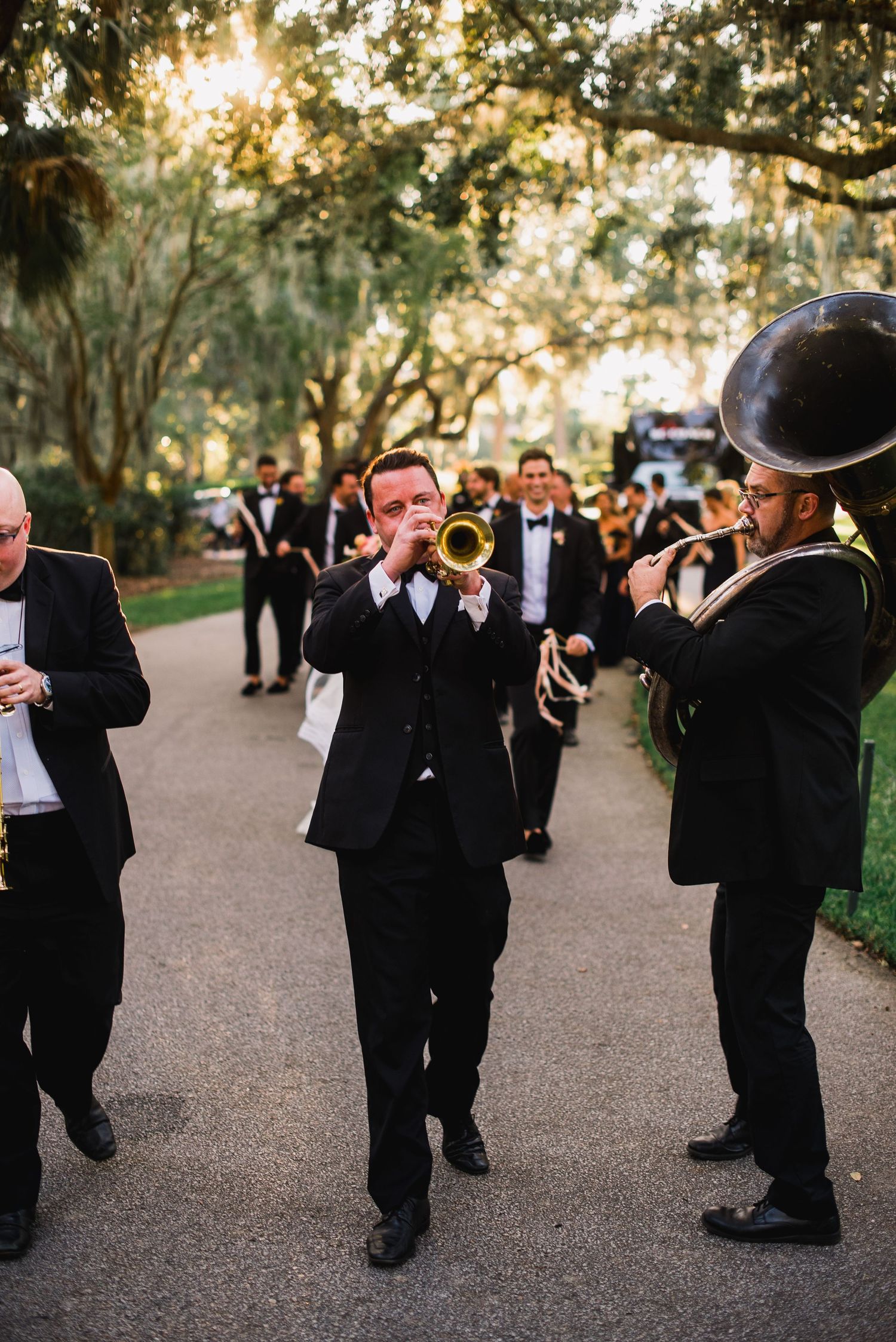 A line of groomsmen in black tuxedos playfully drinking from flasks in a sunlit outdoor setting.
