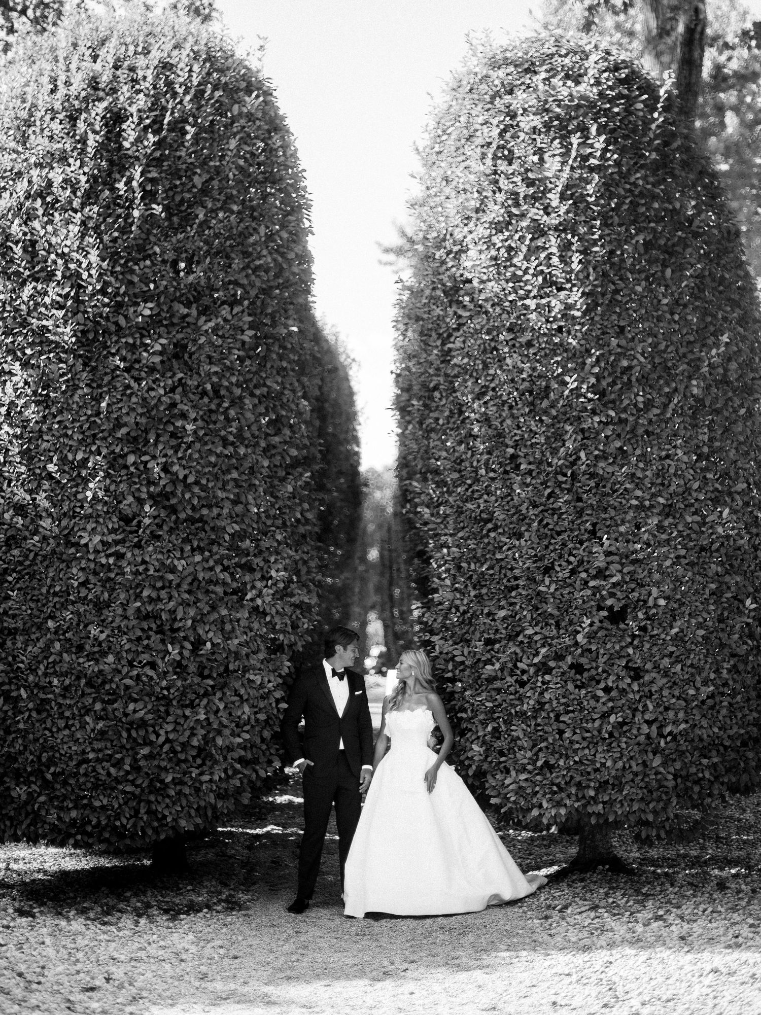 A black and white photo of a couple standing between two tall manicured hedges in a formal garden setting.