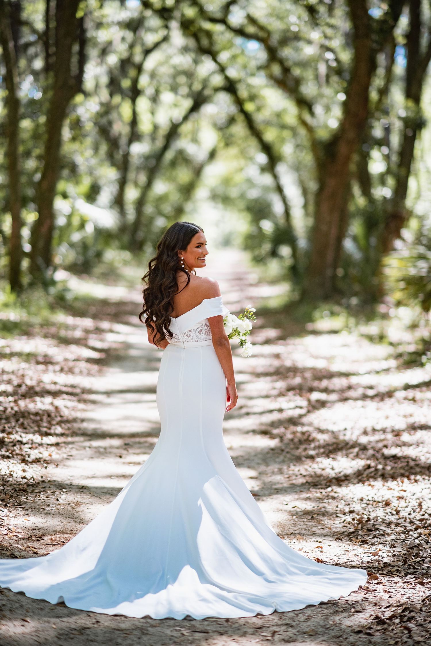 A bride in a flowing white wedding dress twirls gracefully on a sunlit forest path creating a magical outdoor portrait series.