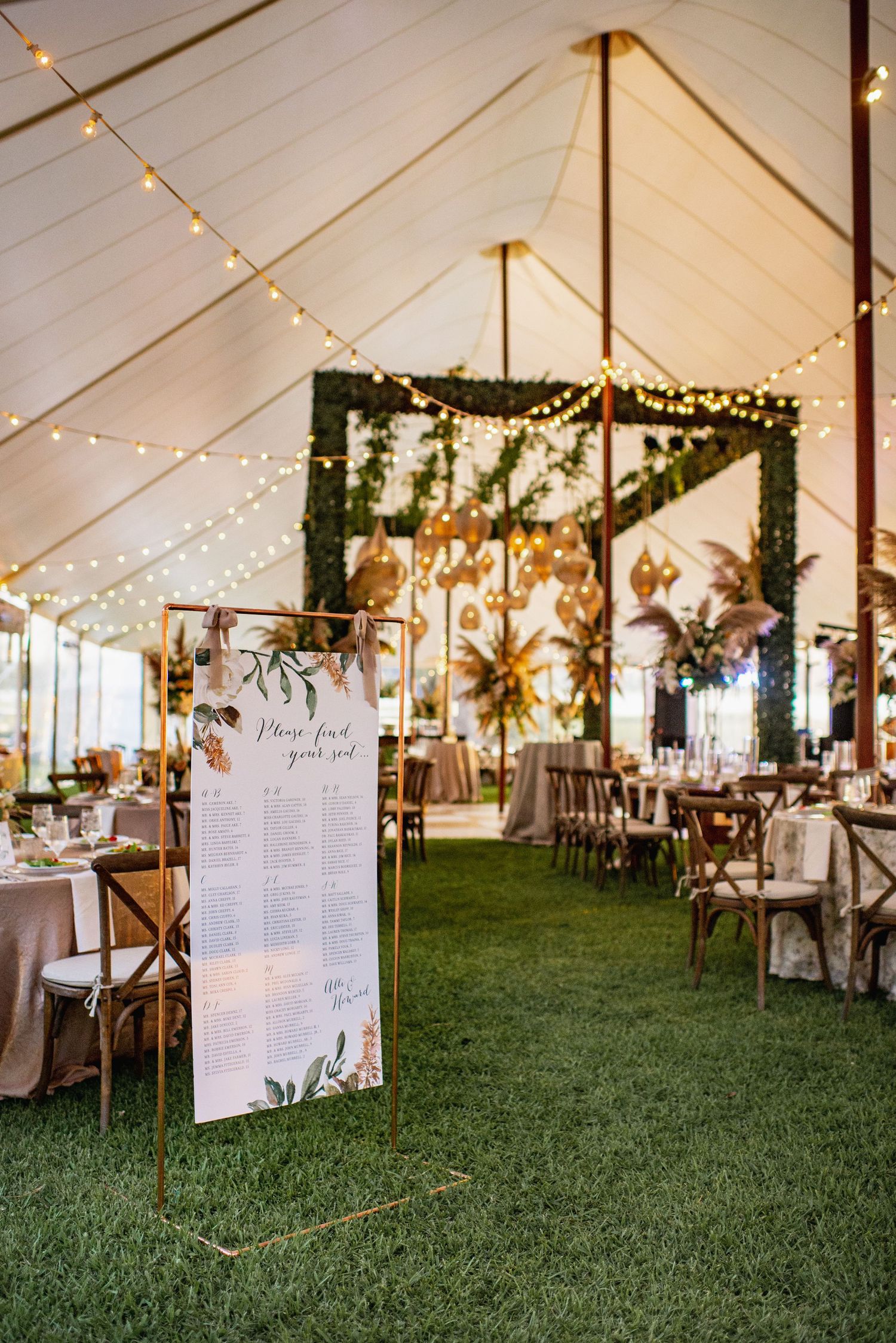 Elegant wedding tent reception setup with string lights, floral decor and a seating chart display on green grass.
